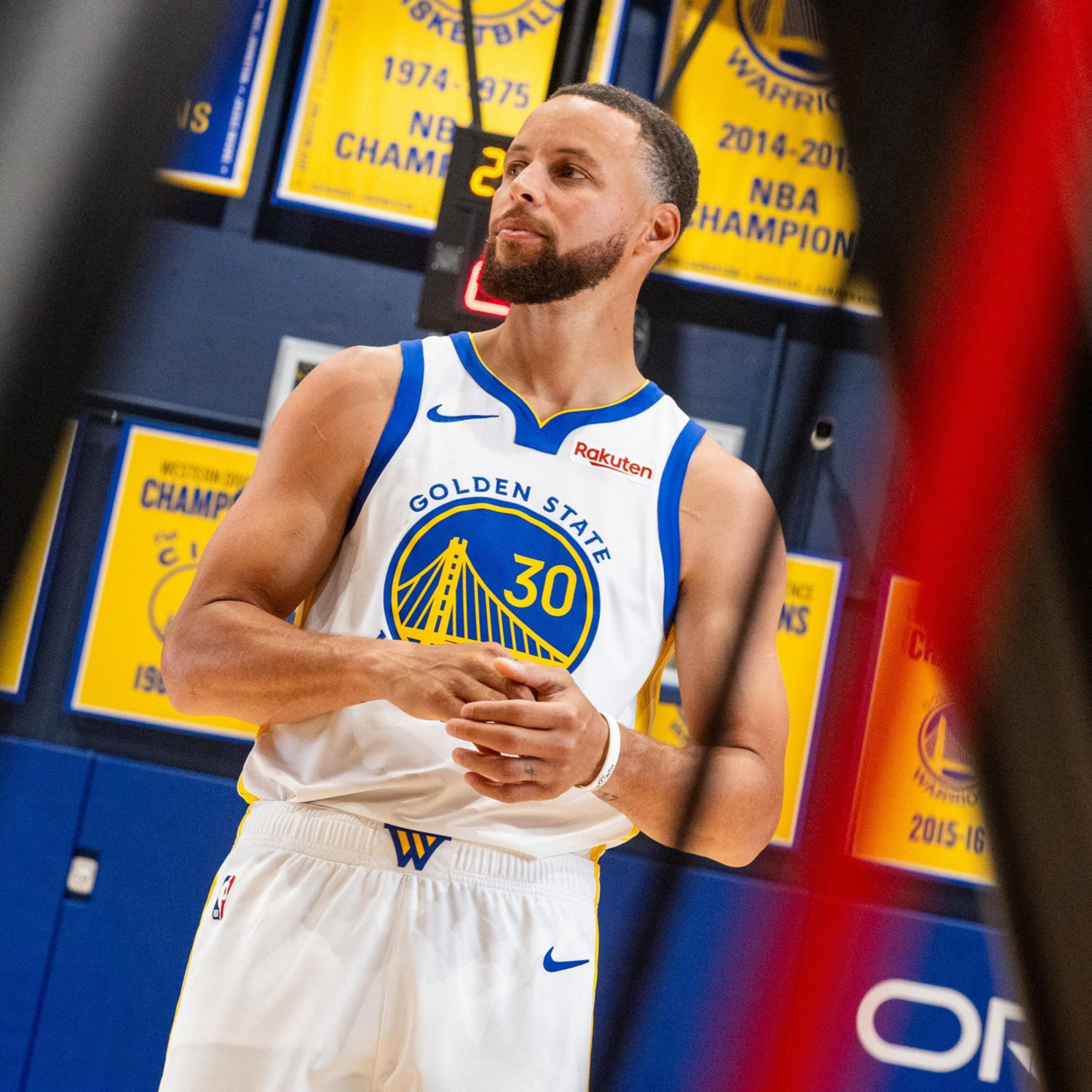 A basketball player in a Golden State Warriors jersey stands in front of championship banners hanging on a gym wall.