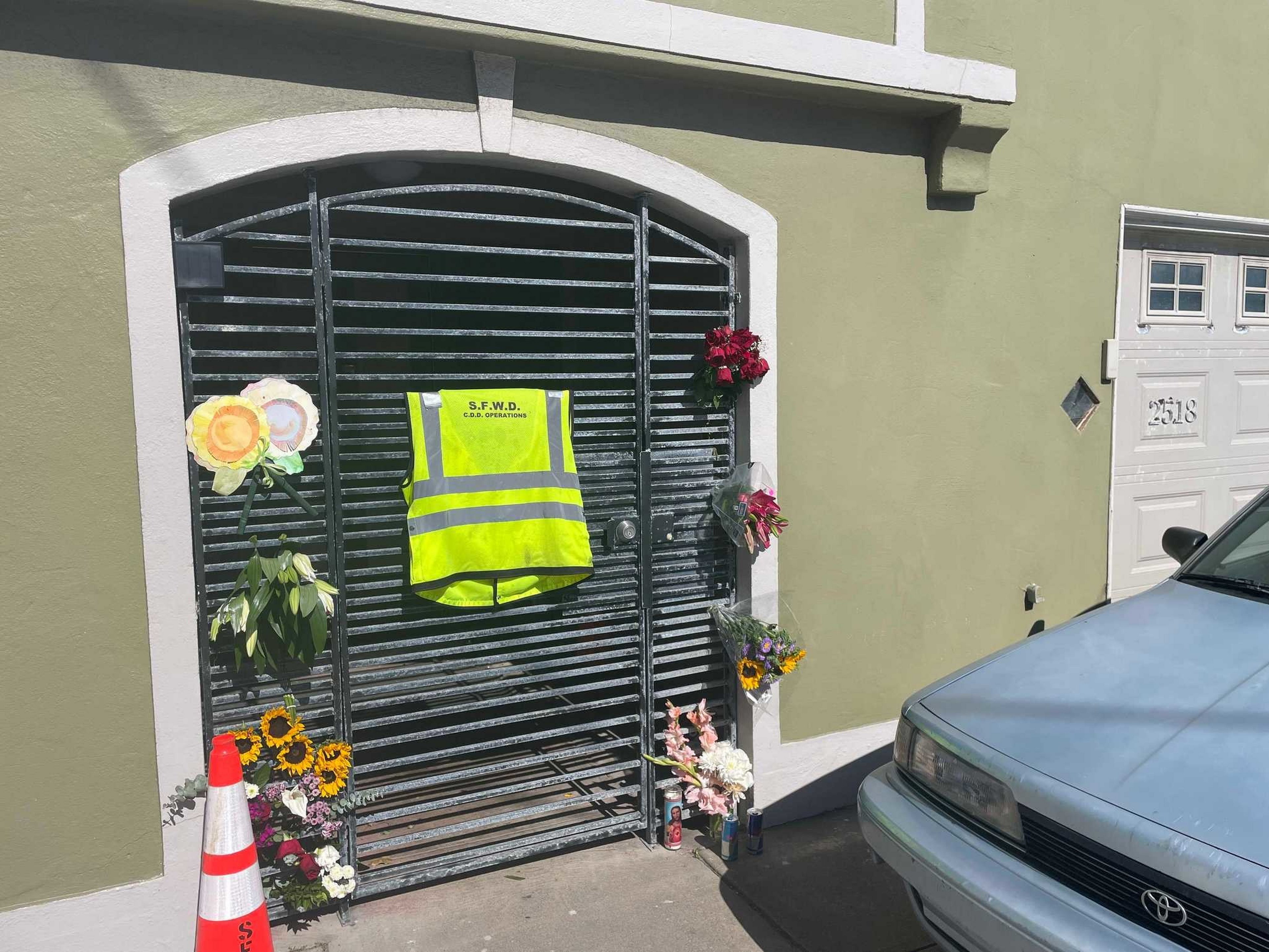 A black metal gate with a yellow reflective vest, surrounded by flowers and a traffic cone, next to a green wall and a blue car.