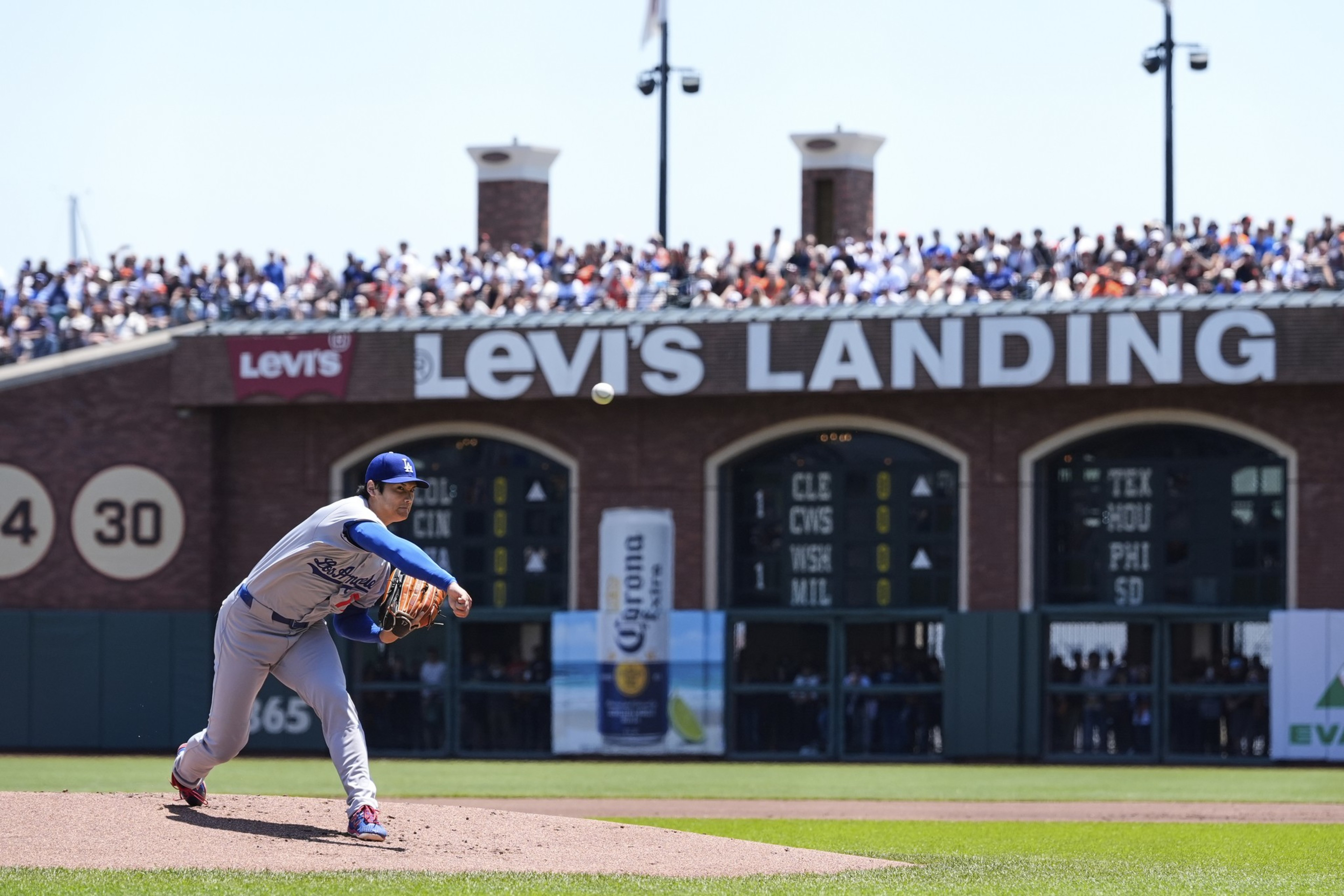 A baseball pitcher in a blue and white uniform throws a pitch on a sunlit field. Behind him, a packed crowd sits under the "Levi's Landing" sign.