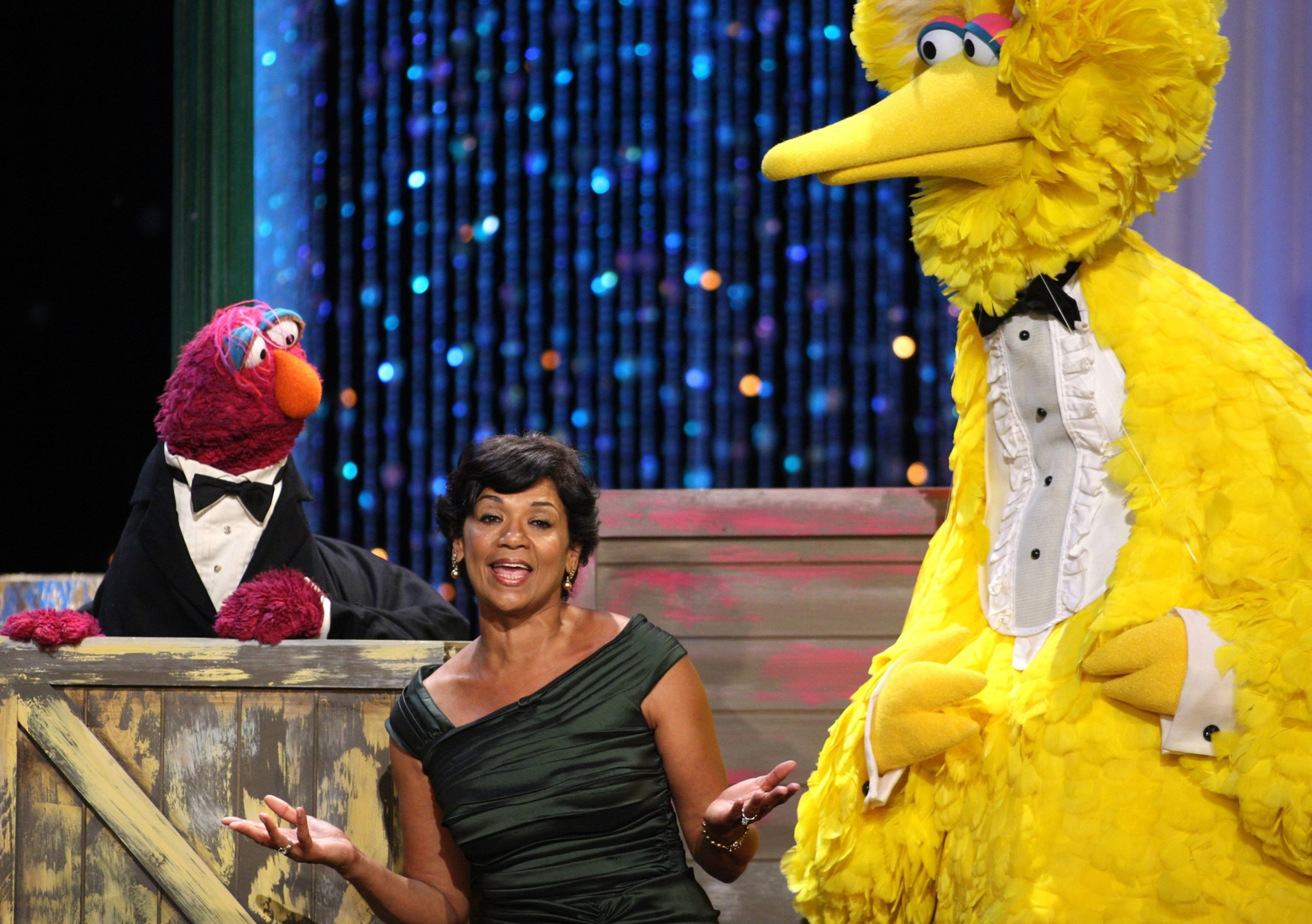 A woman in a dark green dress stands between two puppets dressed in tuxedos, one large yellow bird and one smaller red creature.