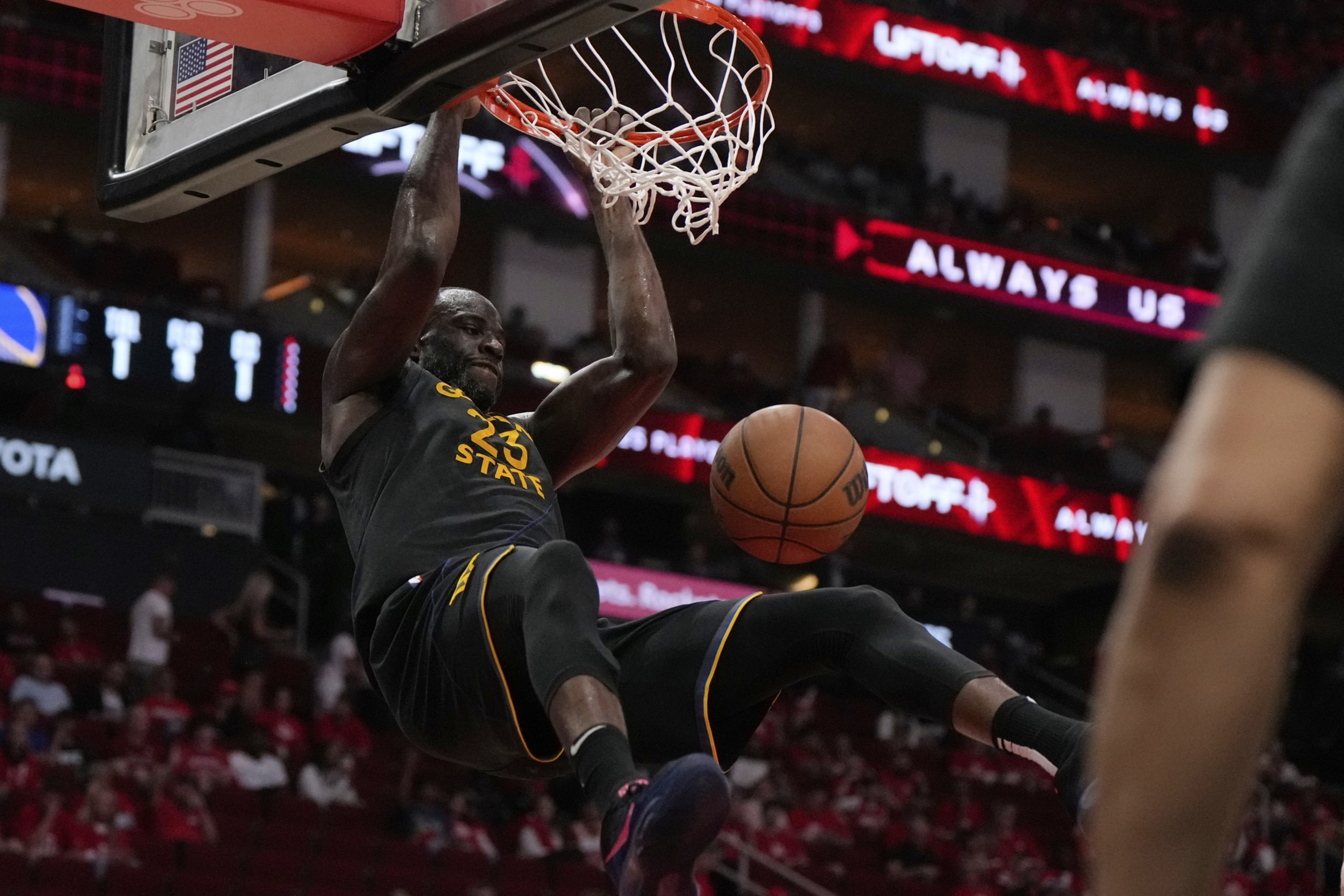 A basketball player in a black "Golden State" jersey hangs from the hoop after a slam dunk, with the ball dropping toward the floor in an indoor arena.