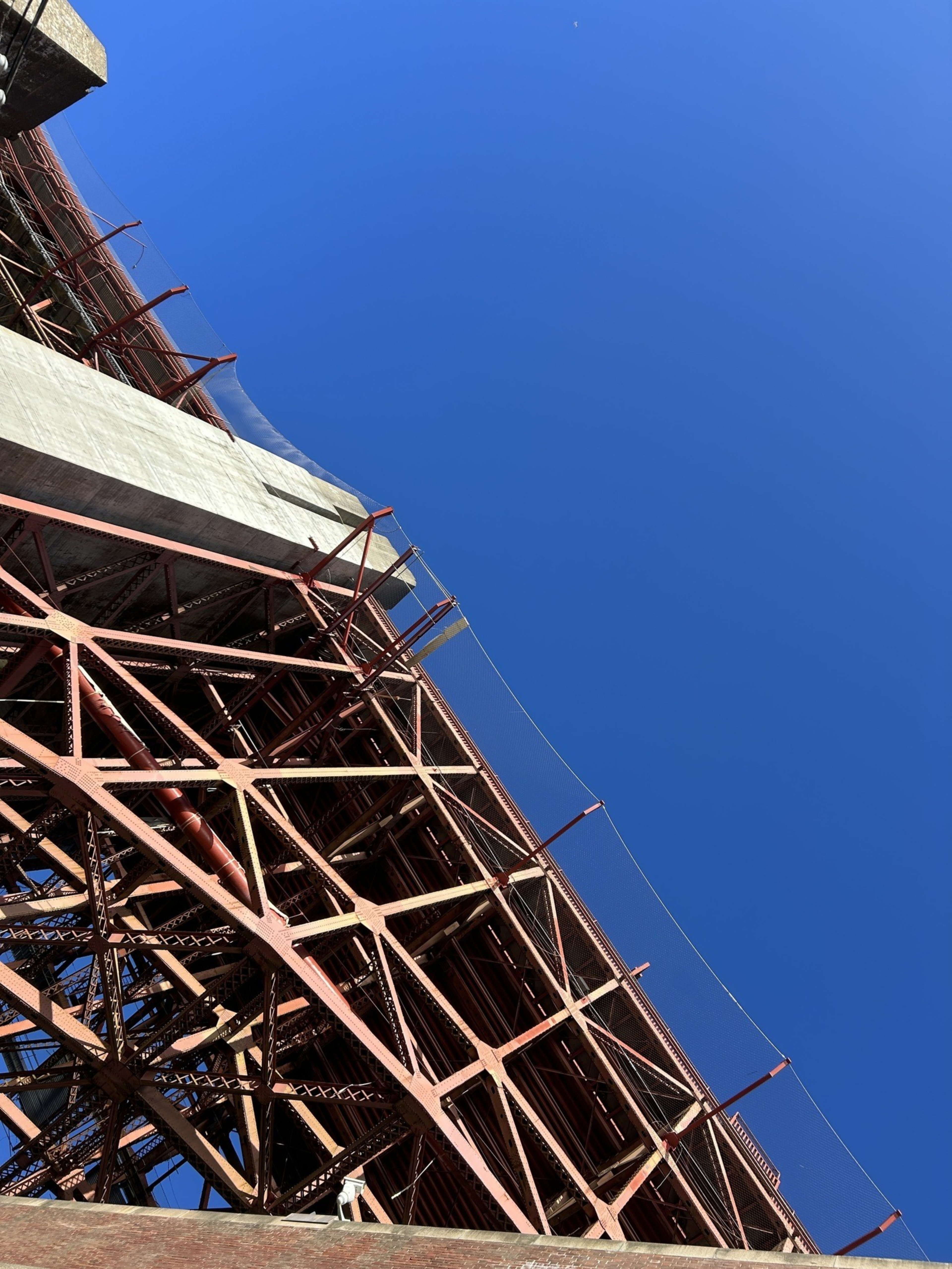 The image shows the underside of a red steel bridge structure against a clear blue sky. Diagonal metal beams and nets are visible, with some concrete elements.