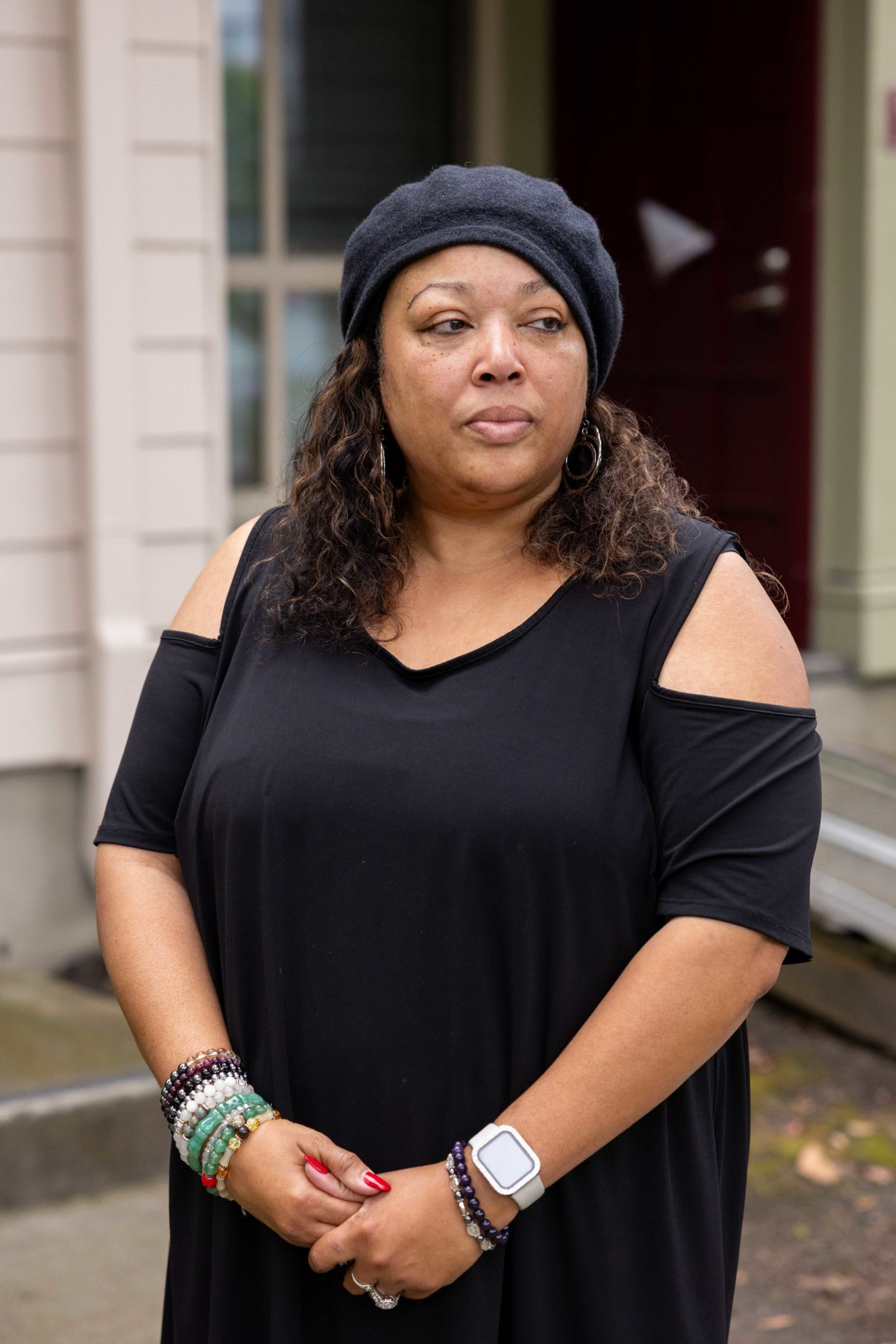 A woman in a black cold-shoulder dress wears a gray beret, hoop earrings, a smartwatch, and multiple beaded bracelets, standing outdoors near a building.
