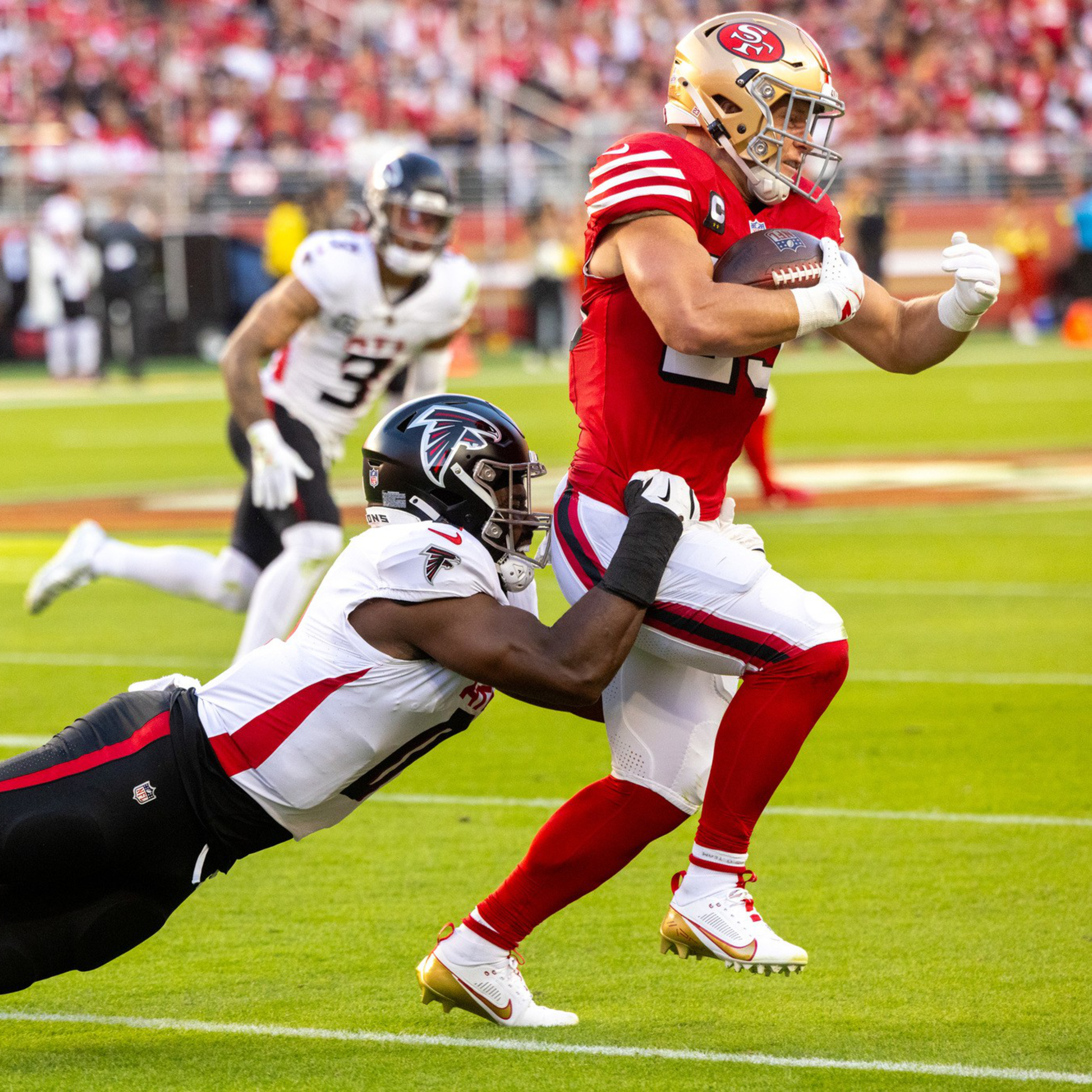 A football player in a red uniform is running with the ball while a player in a white uniform dives, grabbing his leg to tackle him.