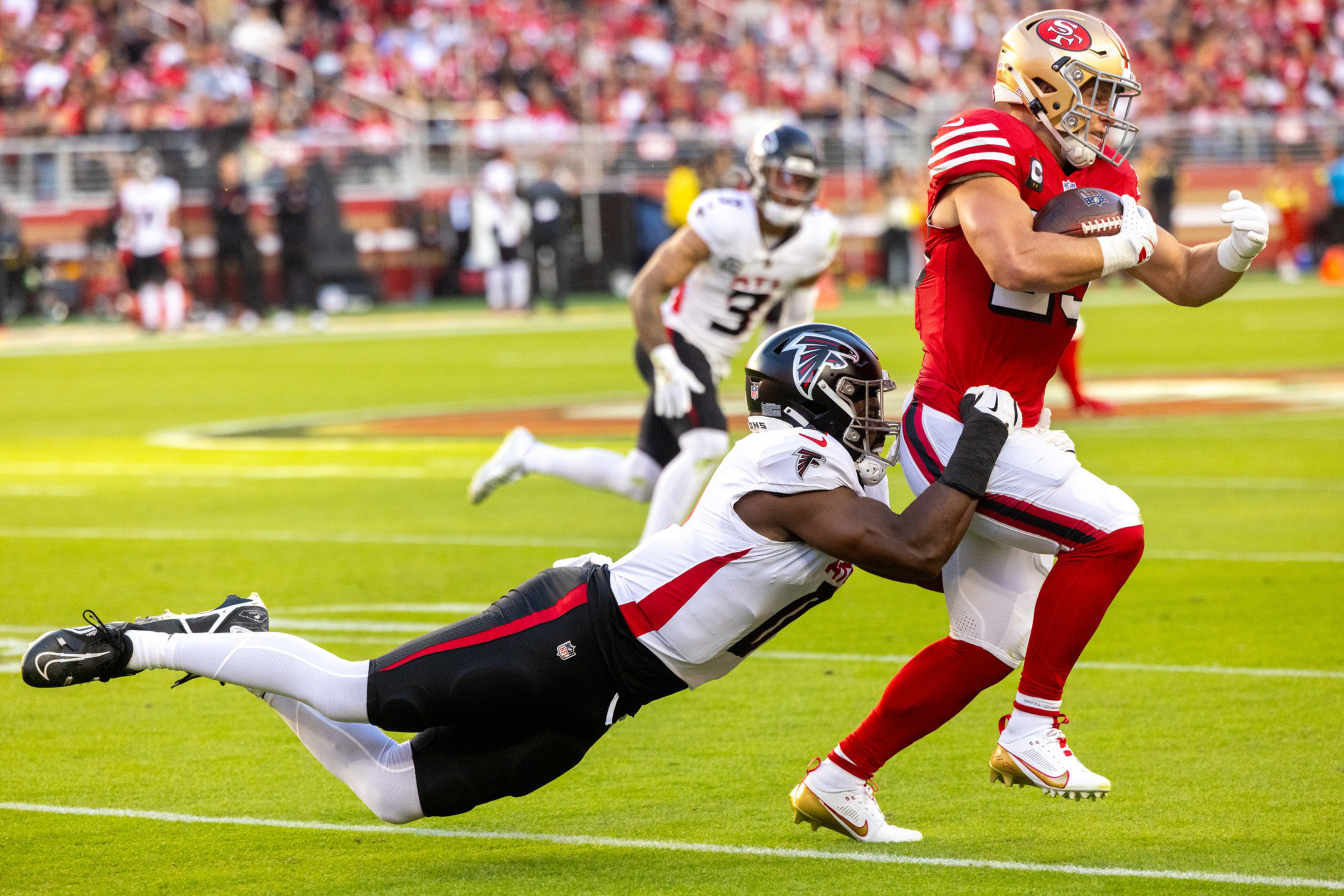 A football player in a red uniform is running with the ball while a player in a white uniform dives, grabbing his leg to tackle him.