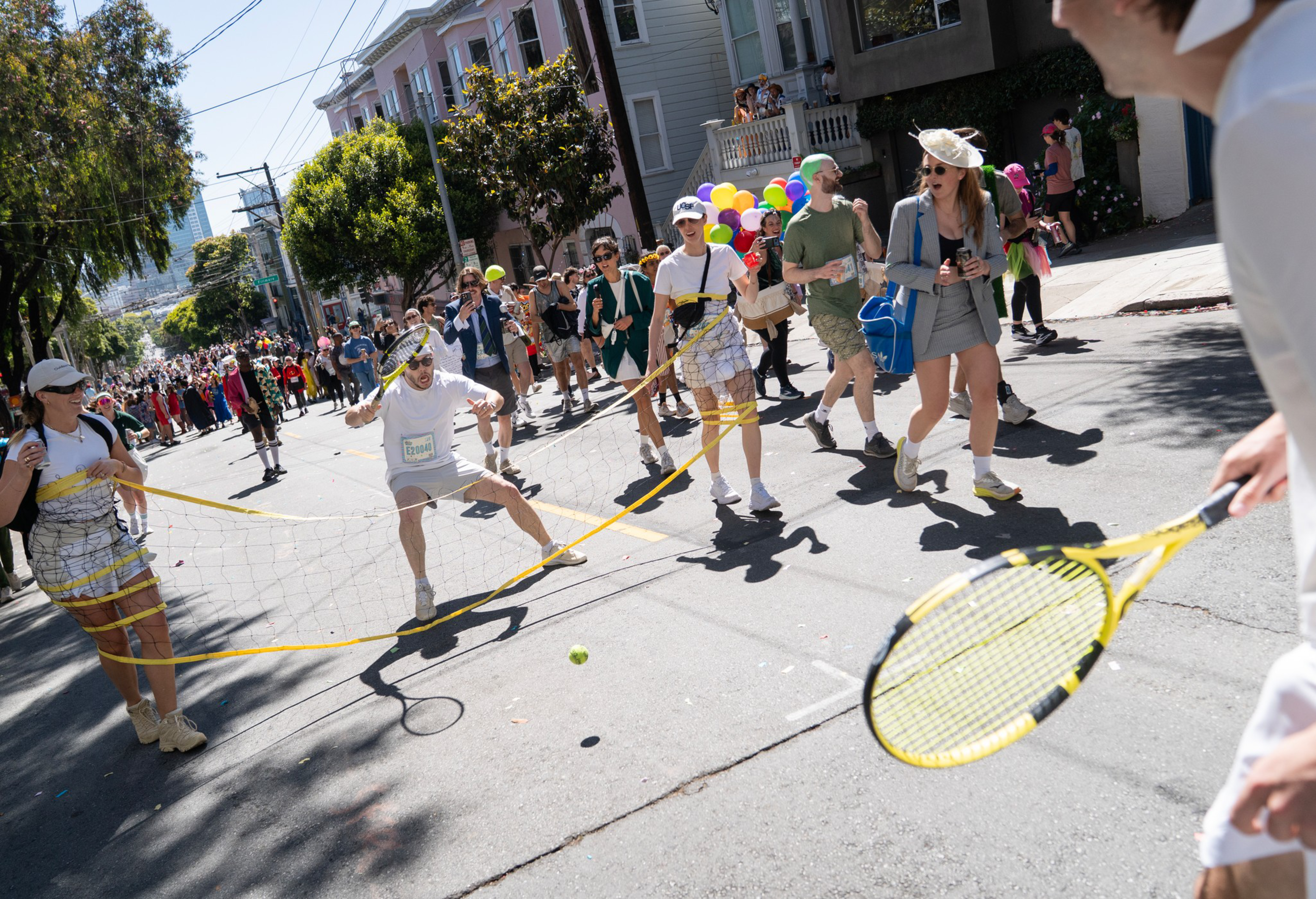 People are playing tennis with a makeshift net on a sunny street, while others watch and walk by, with colorful balloons in the background.