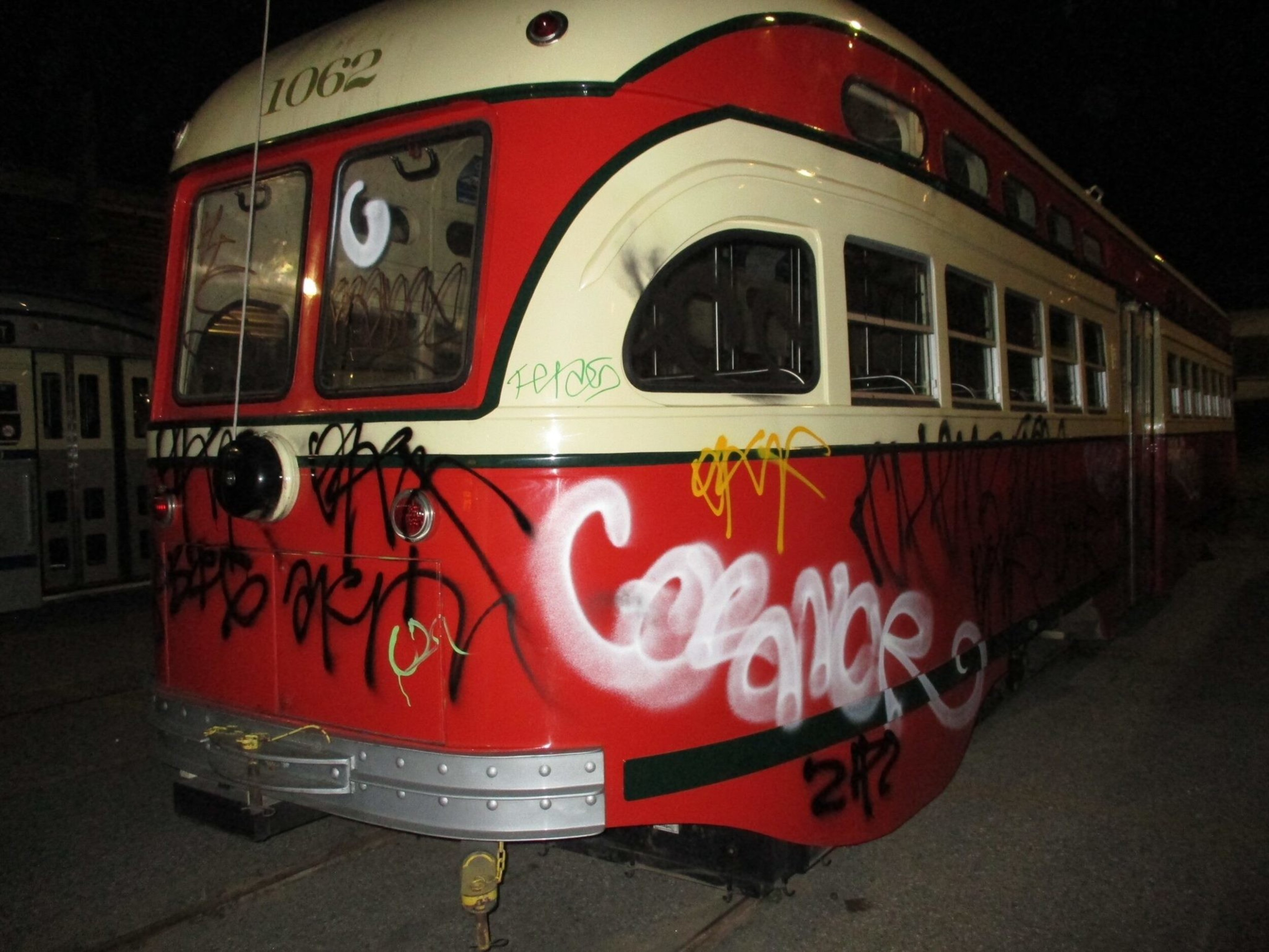 A vintage red and cream streetcar marked 1062 is covered in various graffiti tags, with prominent white and yellow spray paint designs, parked at night.