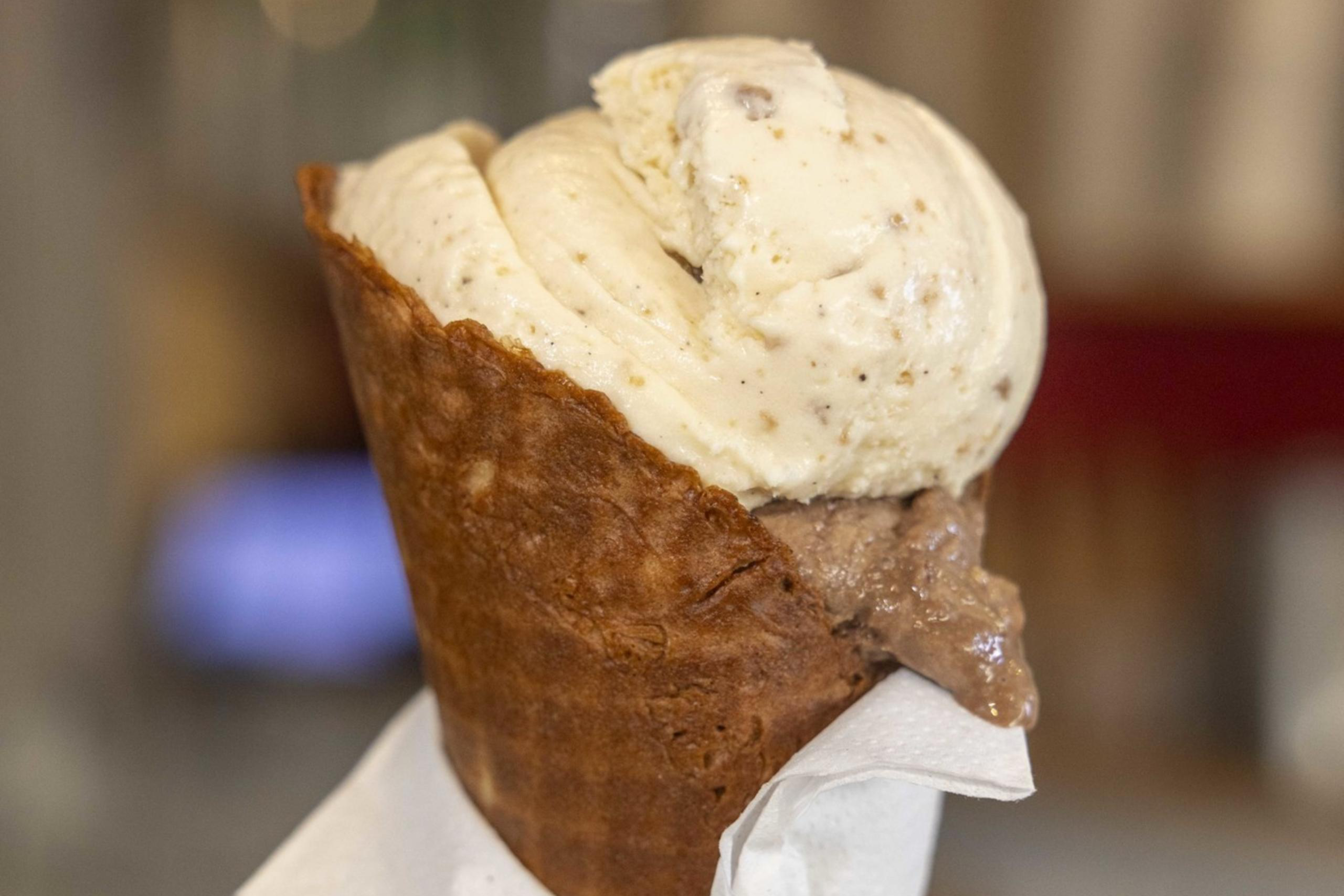 A scoop of creamy vanilla and chocolate ice cream sits in a brown cone wrapped in a white napkin, held upright on a wooden stand.