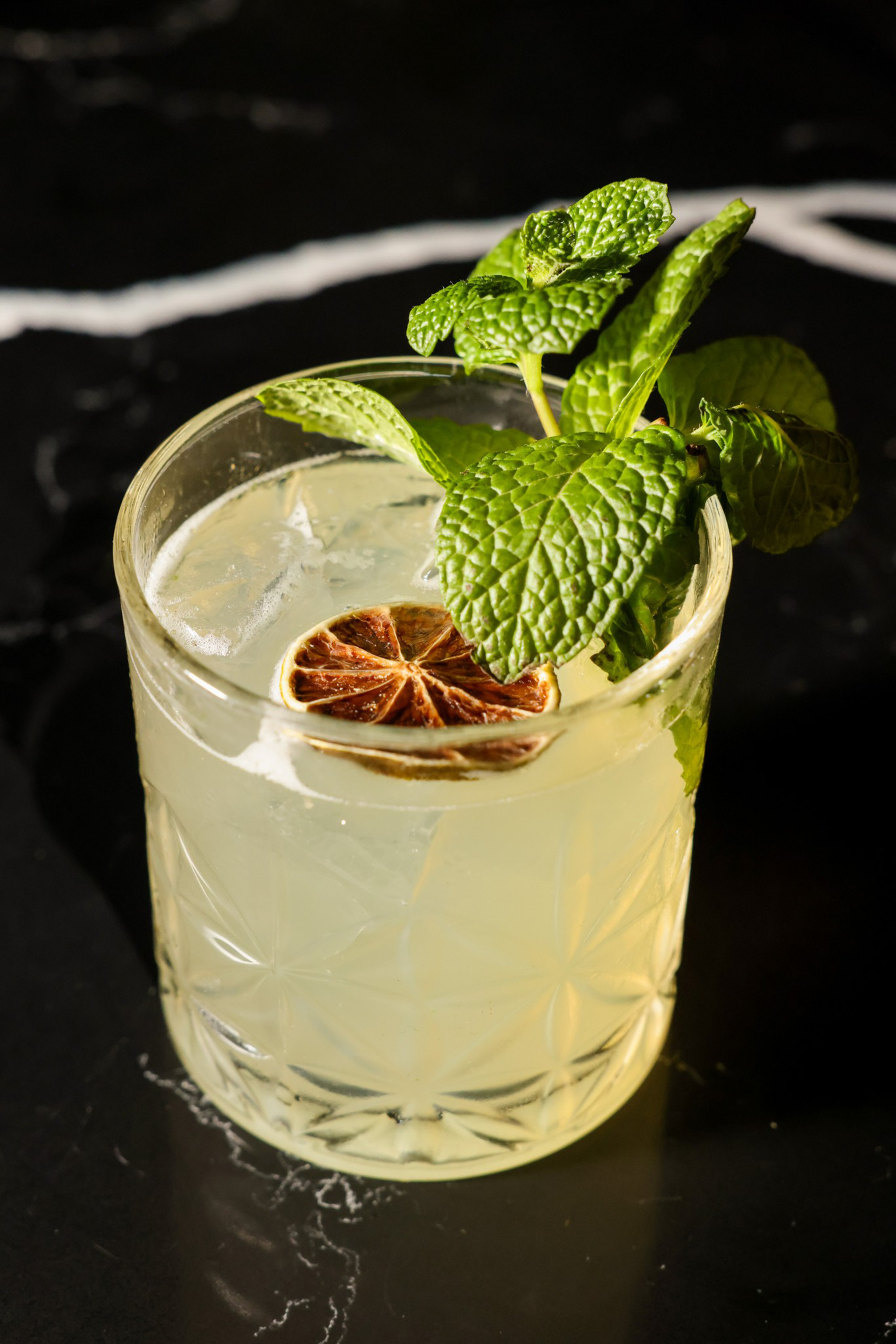 A glass of light yellow cocktail on a dark surface, garnished with a dried citrus slice and fresh mint leaves. Ice cubes are visible in the drink.