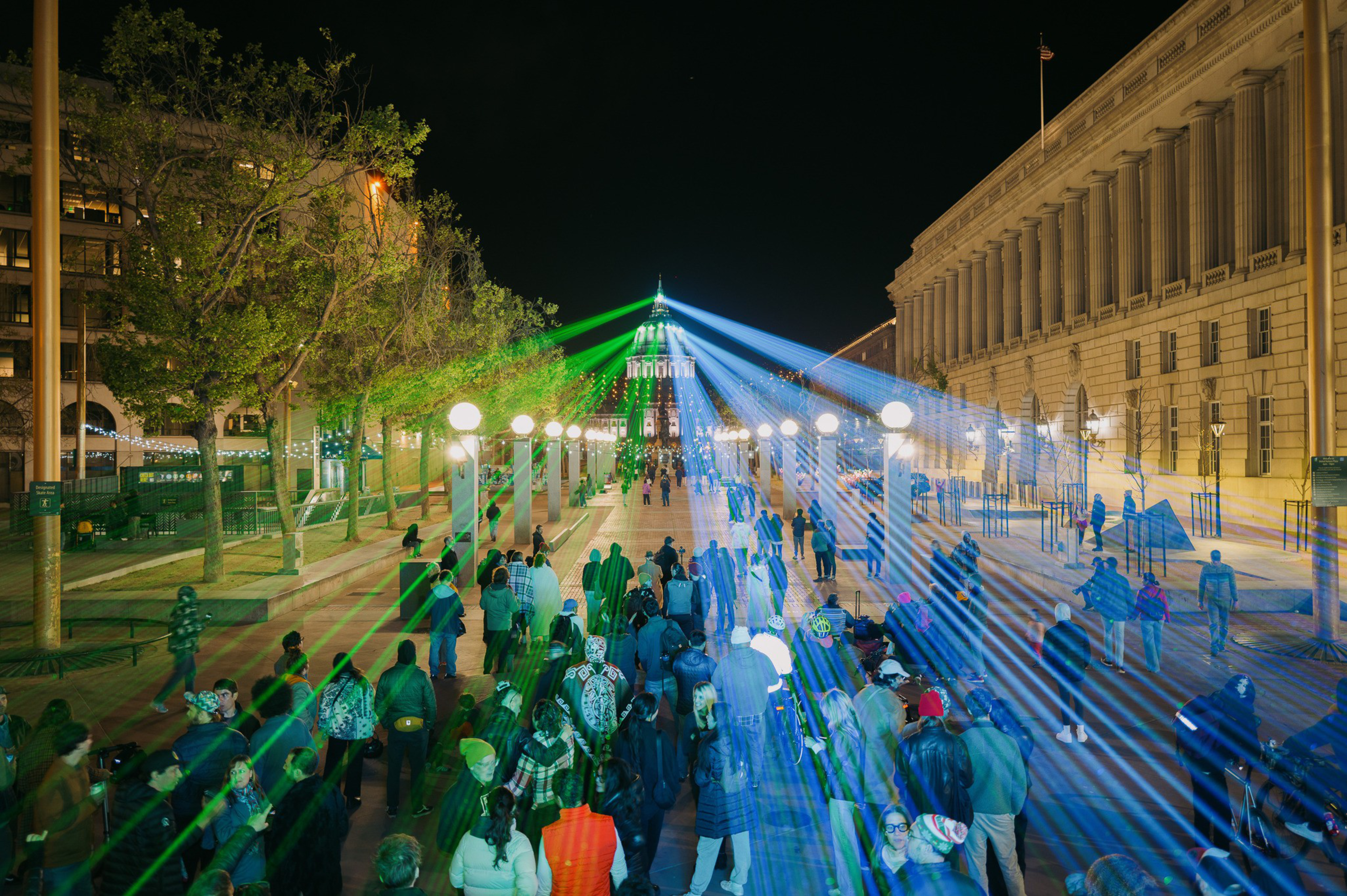 A crowd gathers at night under green and blue laser lights projected towards a distant, illuminated domed building, flanked by trees and grand edifices.