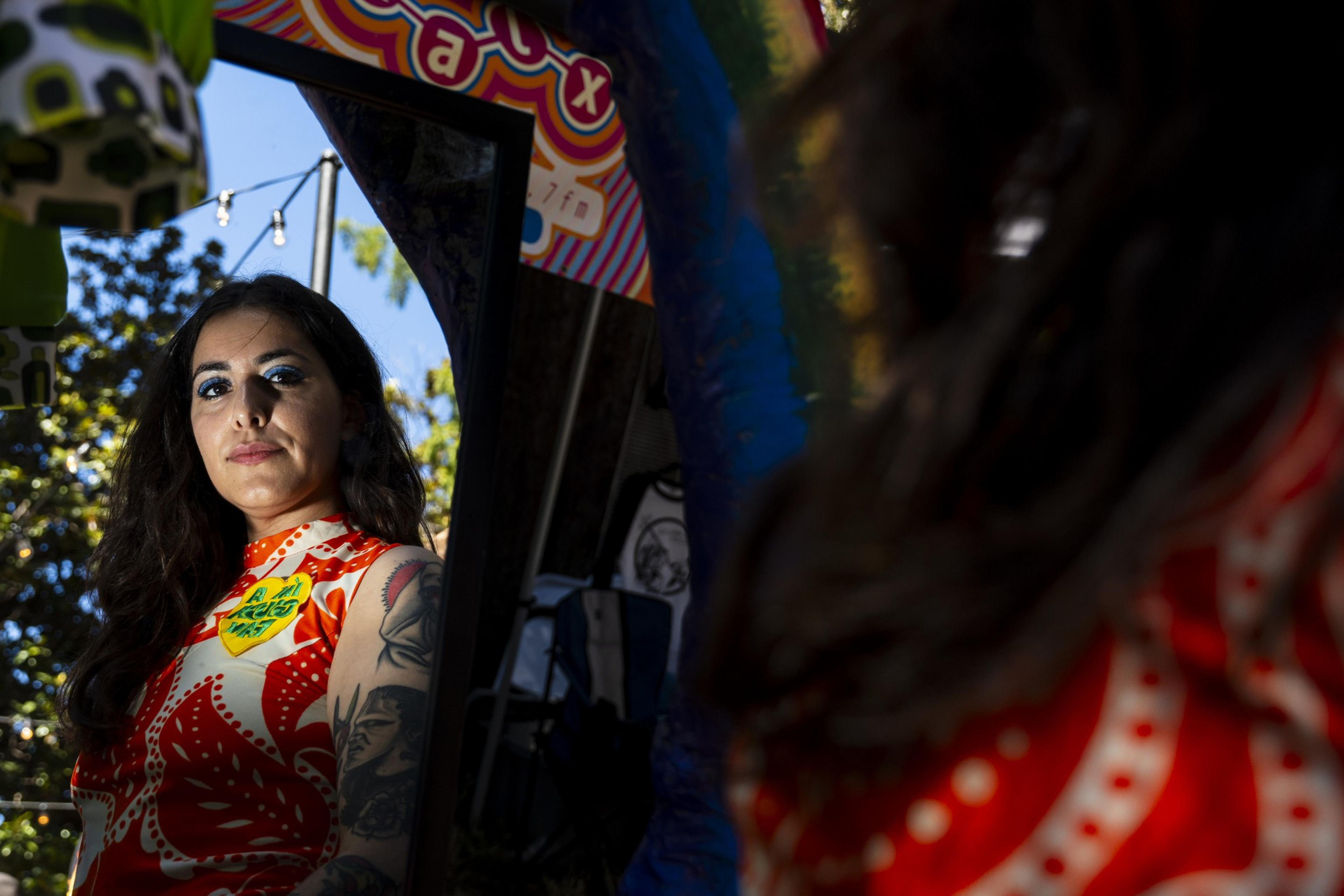 A woman with bold blue eye makeup and tattoos is gazing into a mirror outdoors, wearing a patterned red and white top. The background is vibrant and colorful.