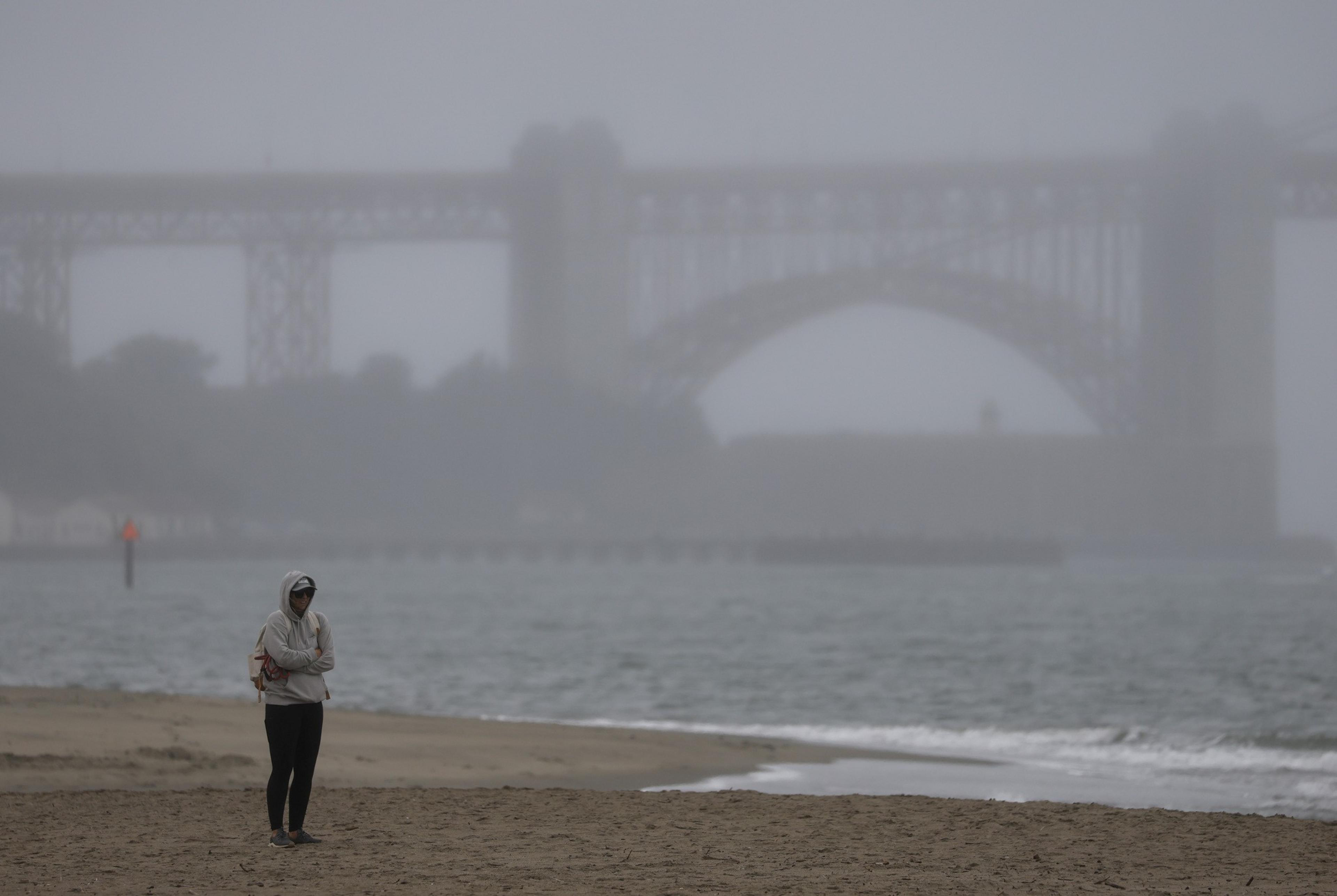 A person in a hoodie walks on a foggy beach near the shoreline. In the background, a large bridge looms faintly in the mist.