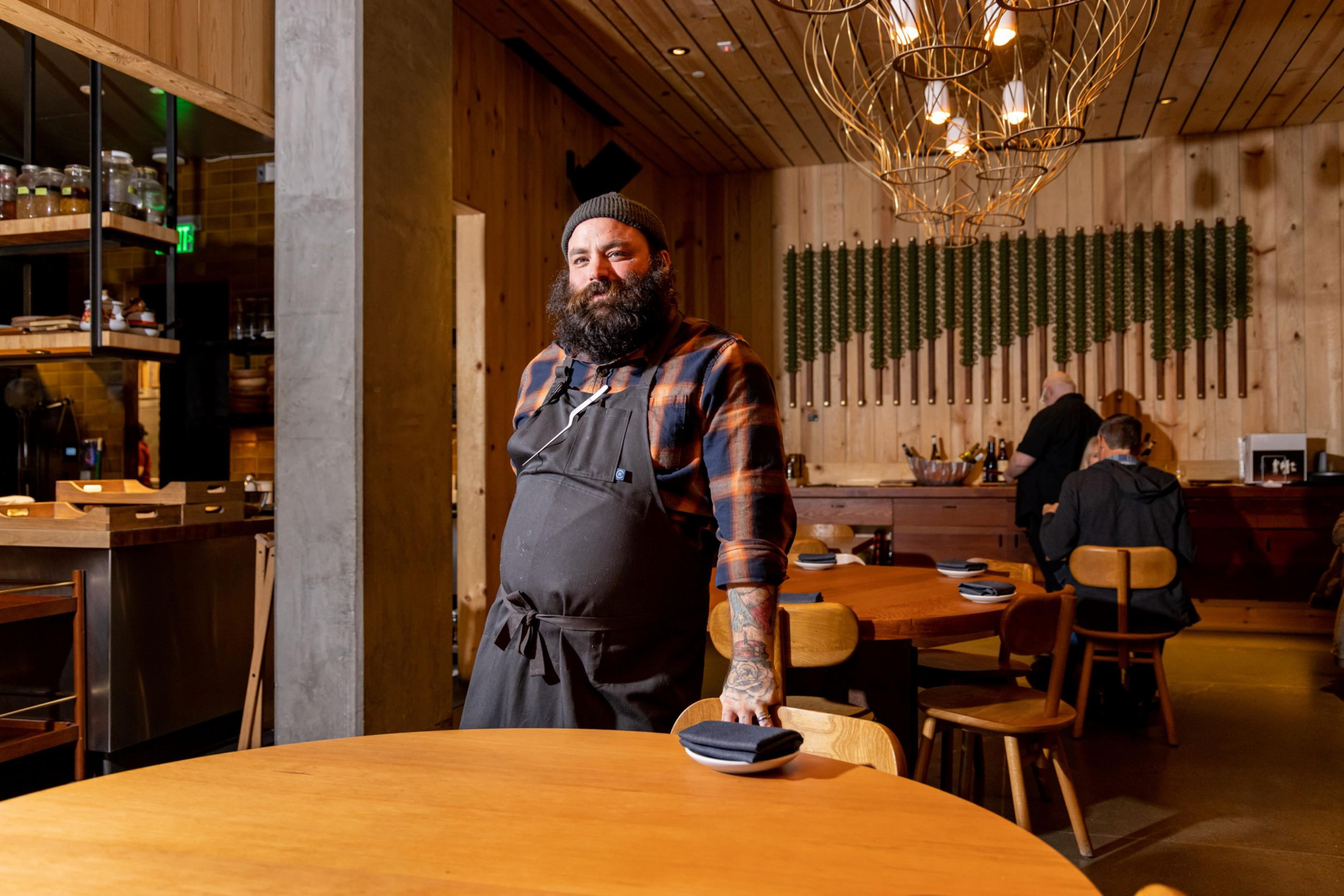 A bearded man in a black apron and plaid shirt stands by a wooden table in a cozy, warmly lit restaurant with wooden interiors.