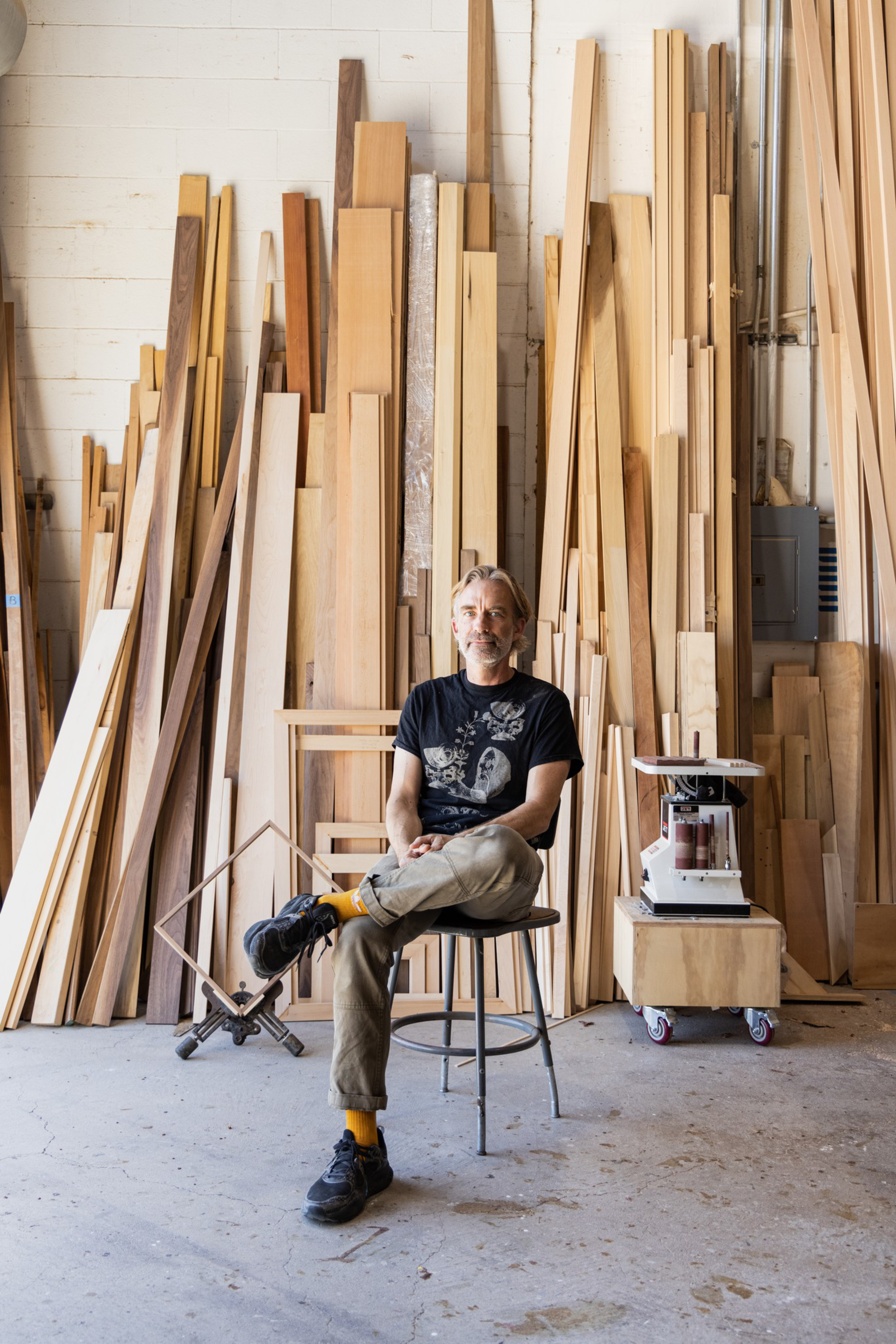 A man with blond hair and glasses sits crossed-legged on a stool in a workshop, surrounded by tall stacks of wooden planks and woodworking tools.