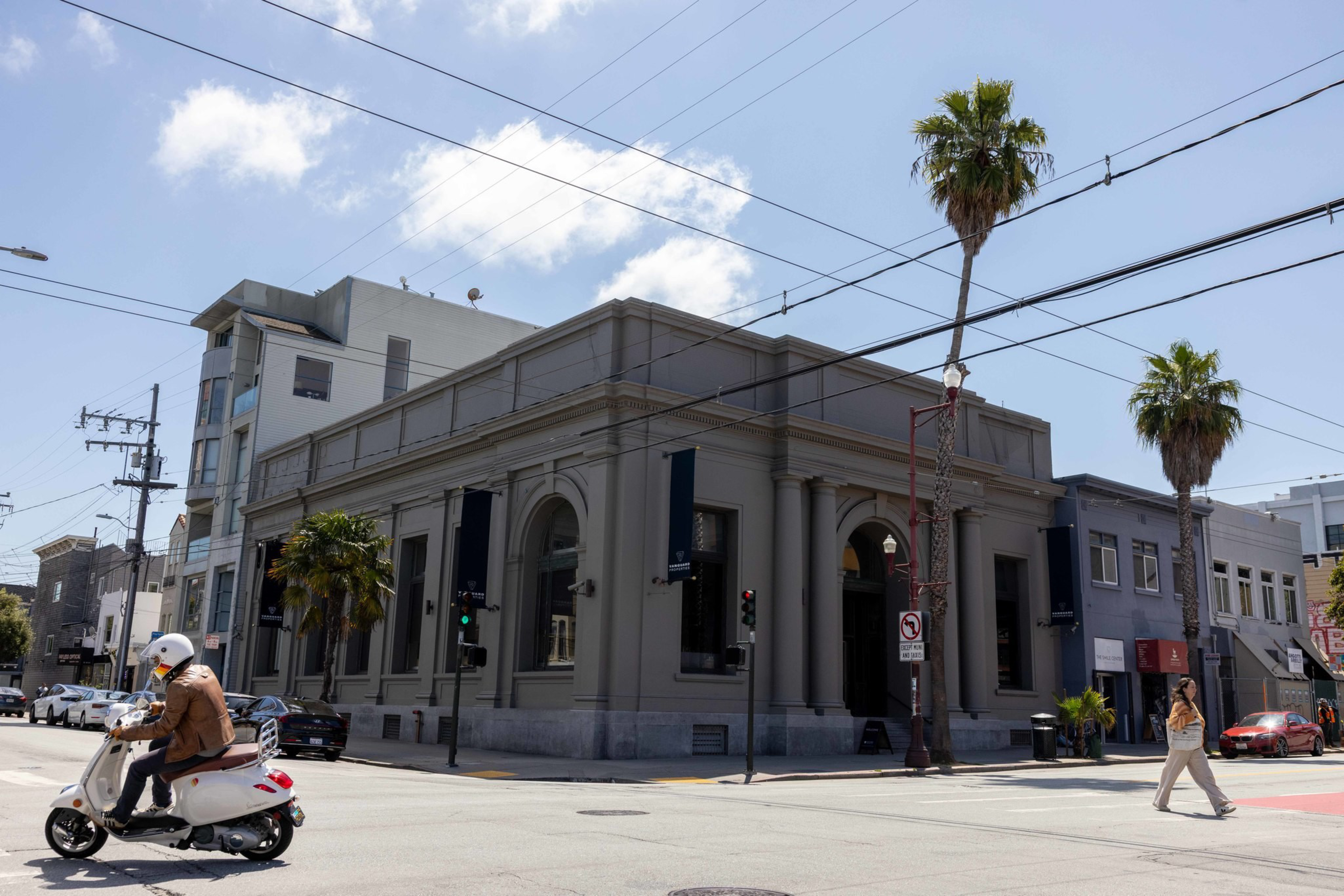 A person on a white scooter waits at an intersection with palm trees, a woman walking, and a large gray building with banners under a partly cloudy sky.