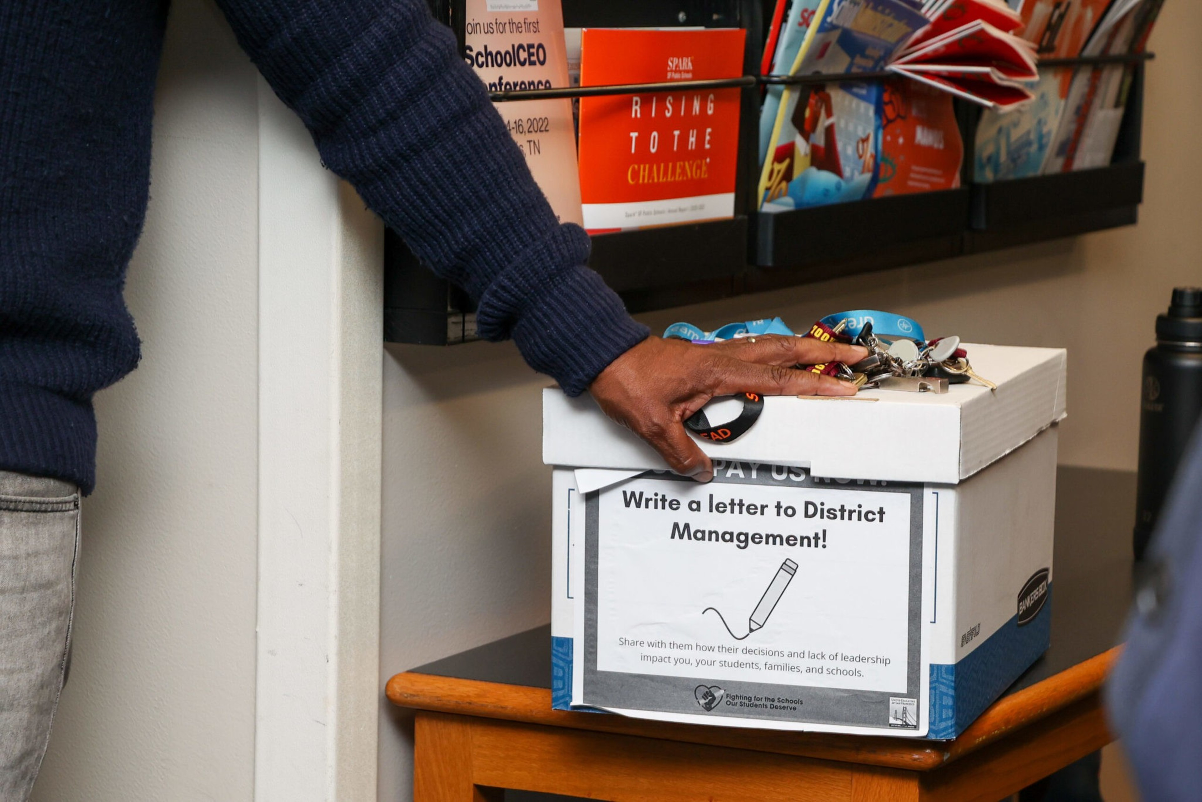A person in a blue sweater places keys on a white box labeled "Write a letter to District Management!" on a wooden table beneath a bookshelf.