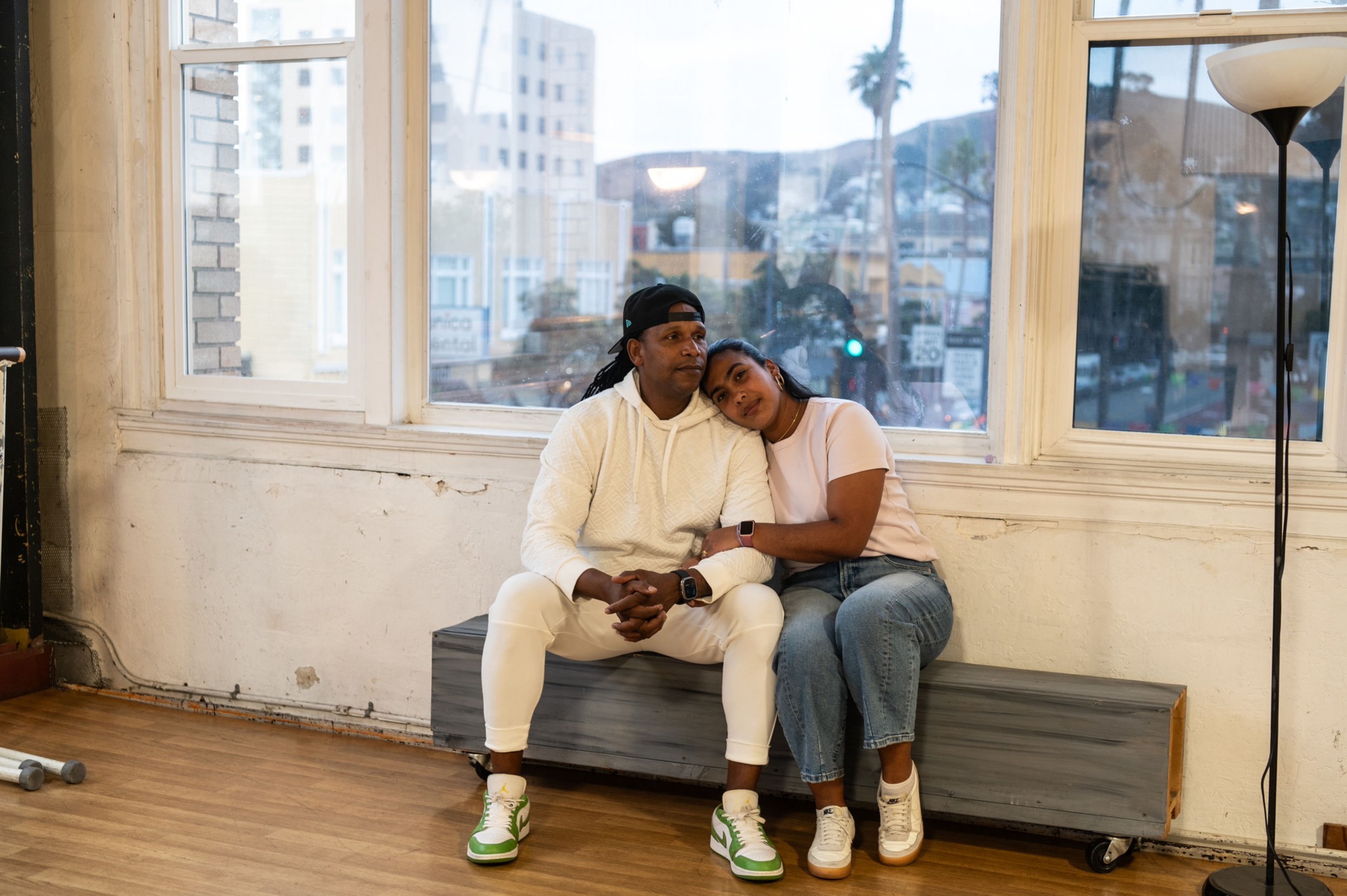 A man in white athletic wear and a woman in a pink shirt and jeans sit closely on a bench, with the woman resting her head on his shoulder.