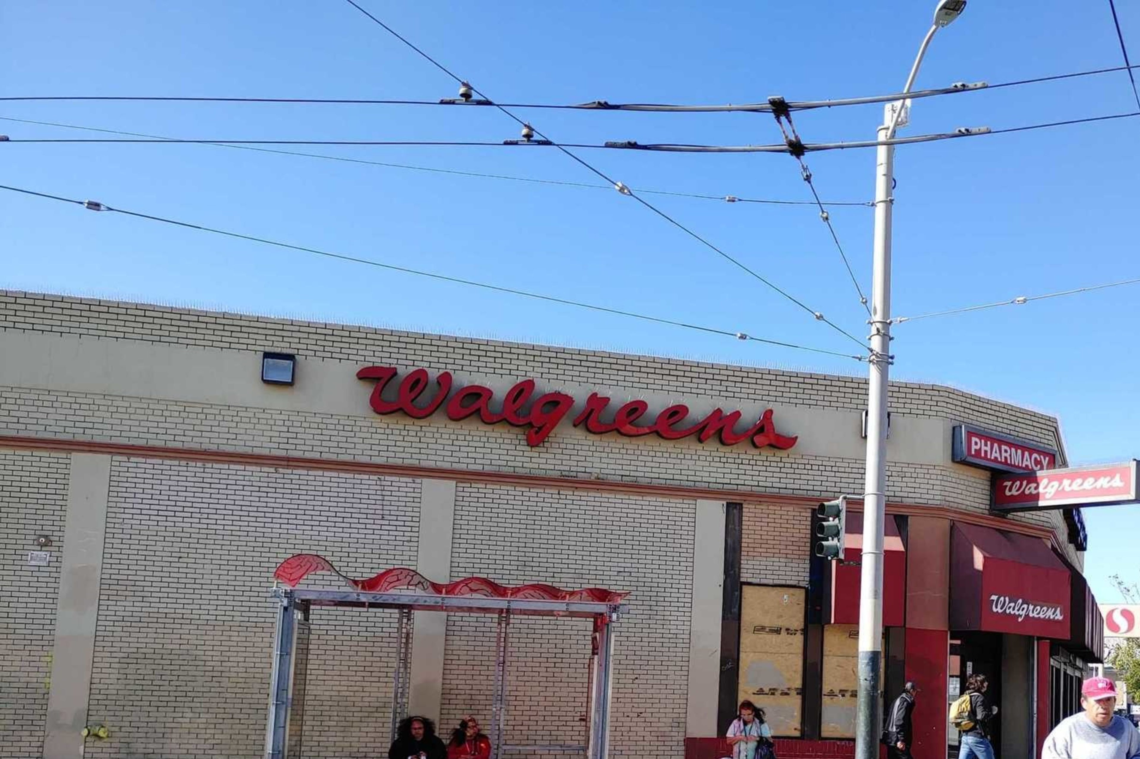 The image shows a Walgreens store on a sunny day, with people walking and sitting at a bus stop. The location is at the corner of a street with a 30th St sign.