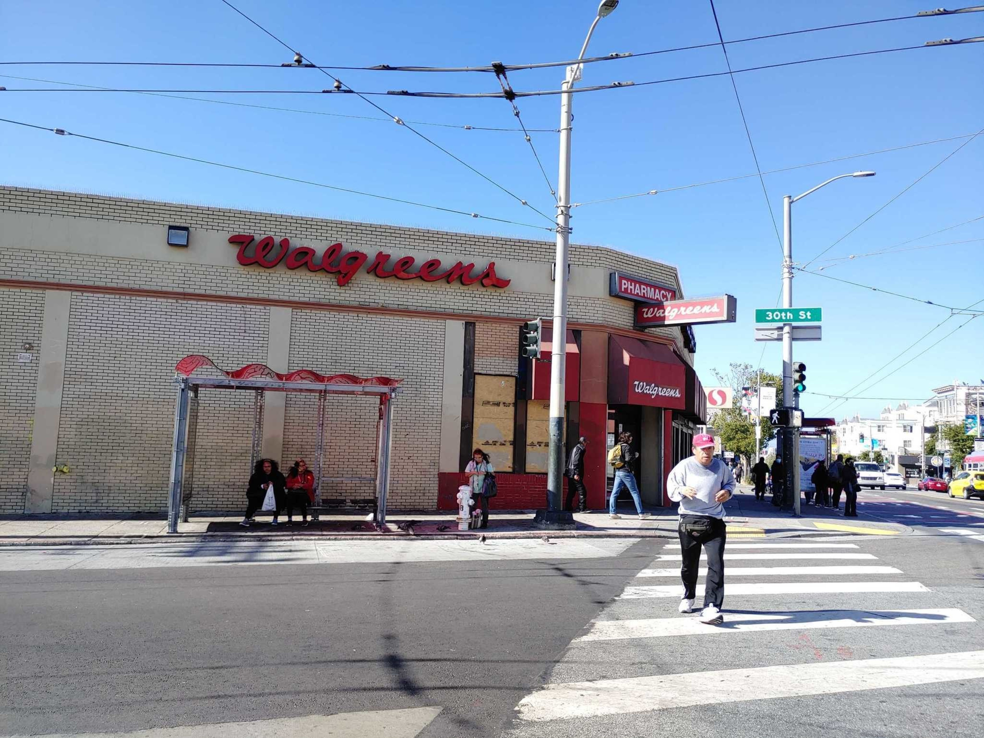 The image shows a Walgreens store on a sunny day, with people walking and sitting at a bus stop. The location is at the corner of a street with a 30th St sign.