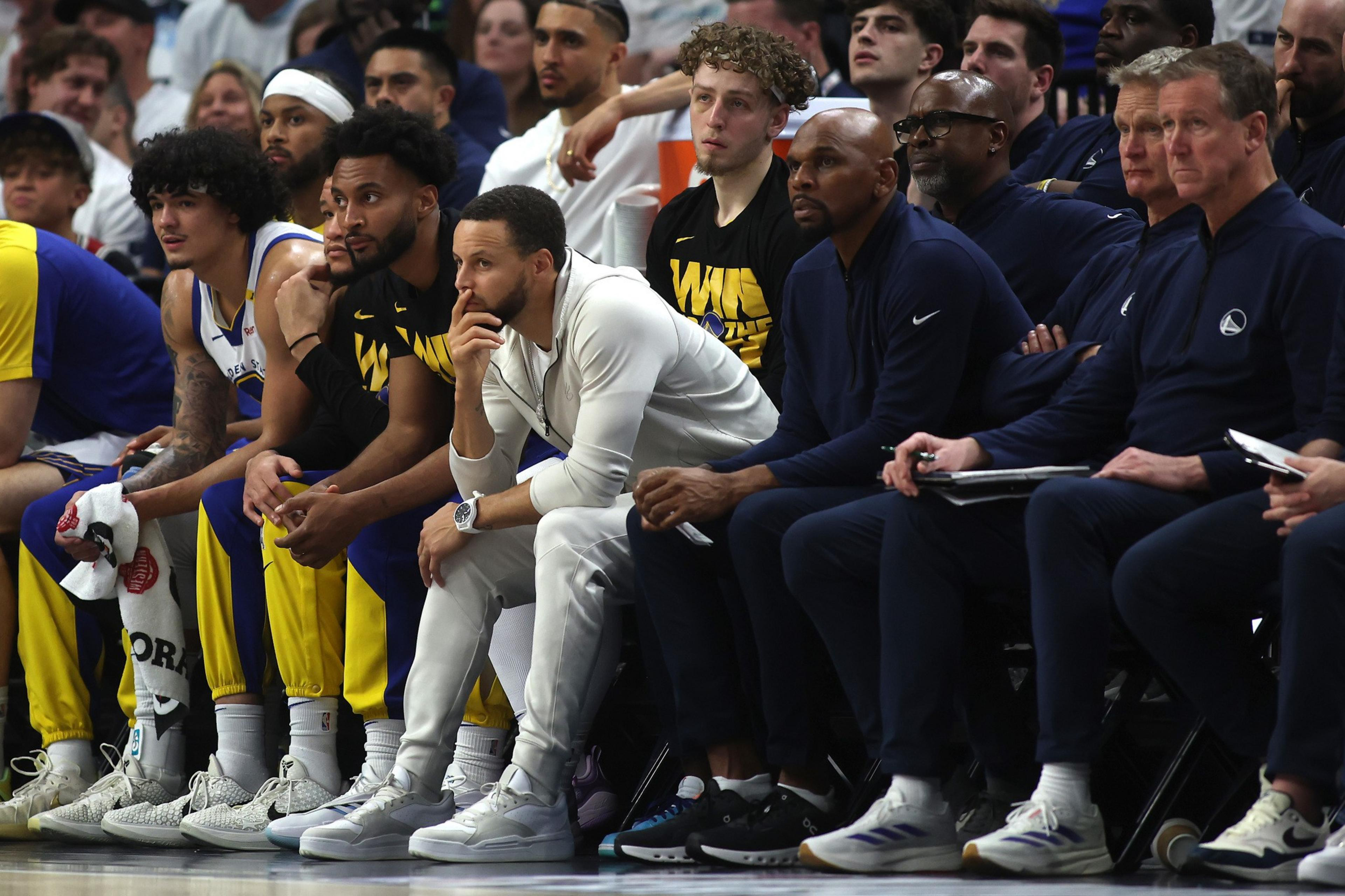 A group of basketball players and coaches sit on a bench. They are focused on the game, with some in team jerseys and others in casual clothes, watching intently.