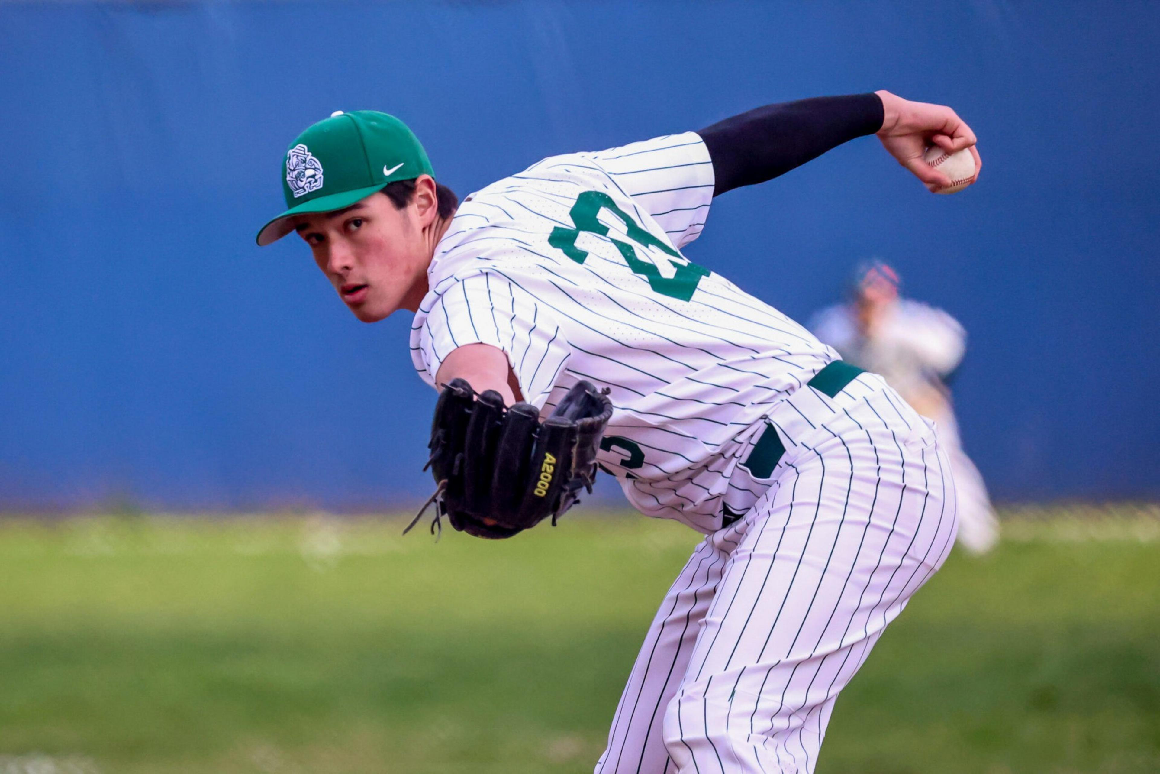 A baseball player in a white pinstriped uniform and green cap is mid-pitch, gripping the ball in his right hand and wearing a black glove on his left.