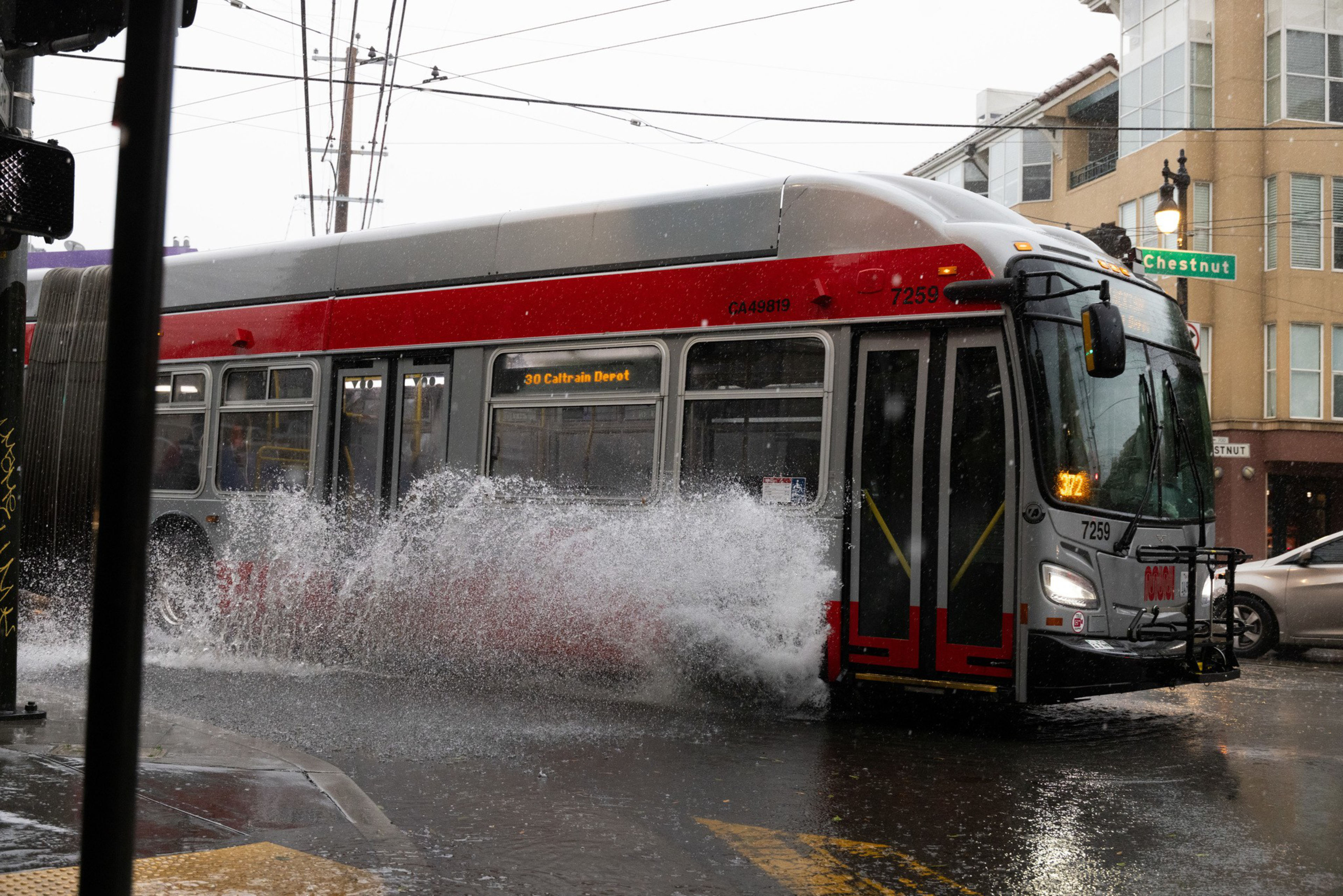 A bus drives through a flooded street, splashing water high into the air. It is overcast and part of a building and street signs are visible in the background.