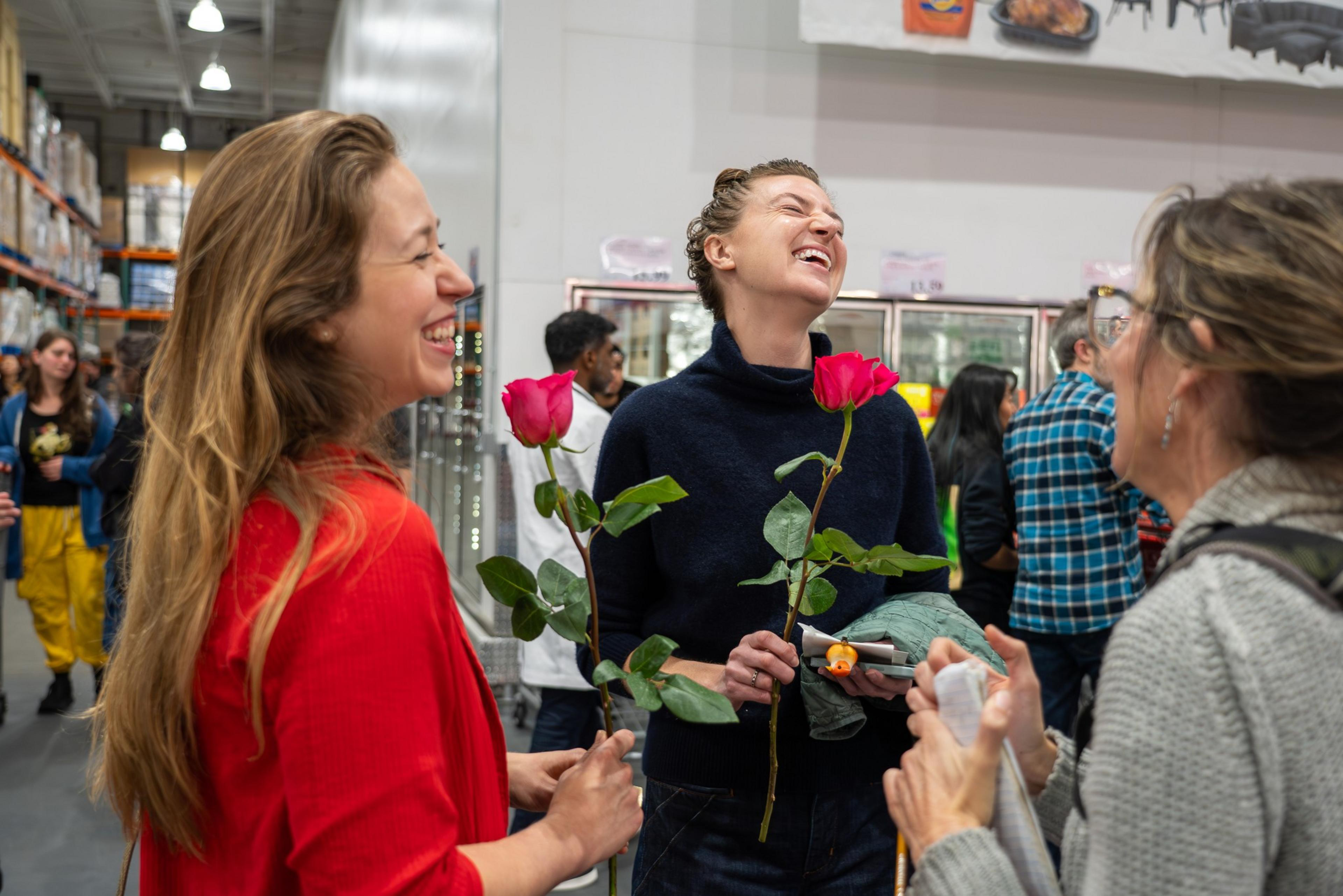 Three women laugh and talk in a store aisle, each holding a pink rose, surrounded by other shoppers and refrigerated displays.
