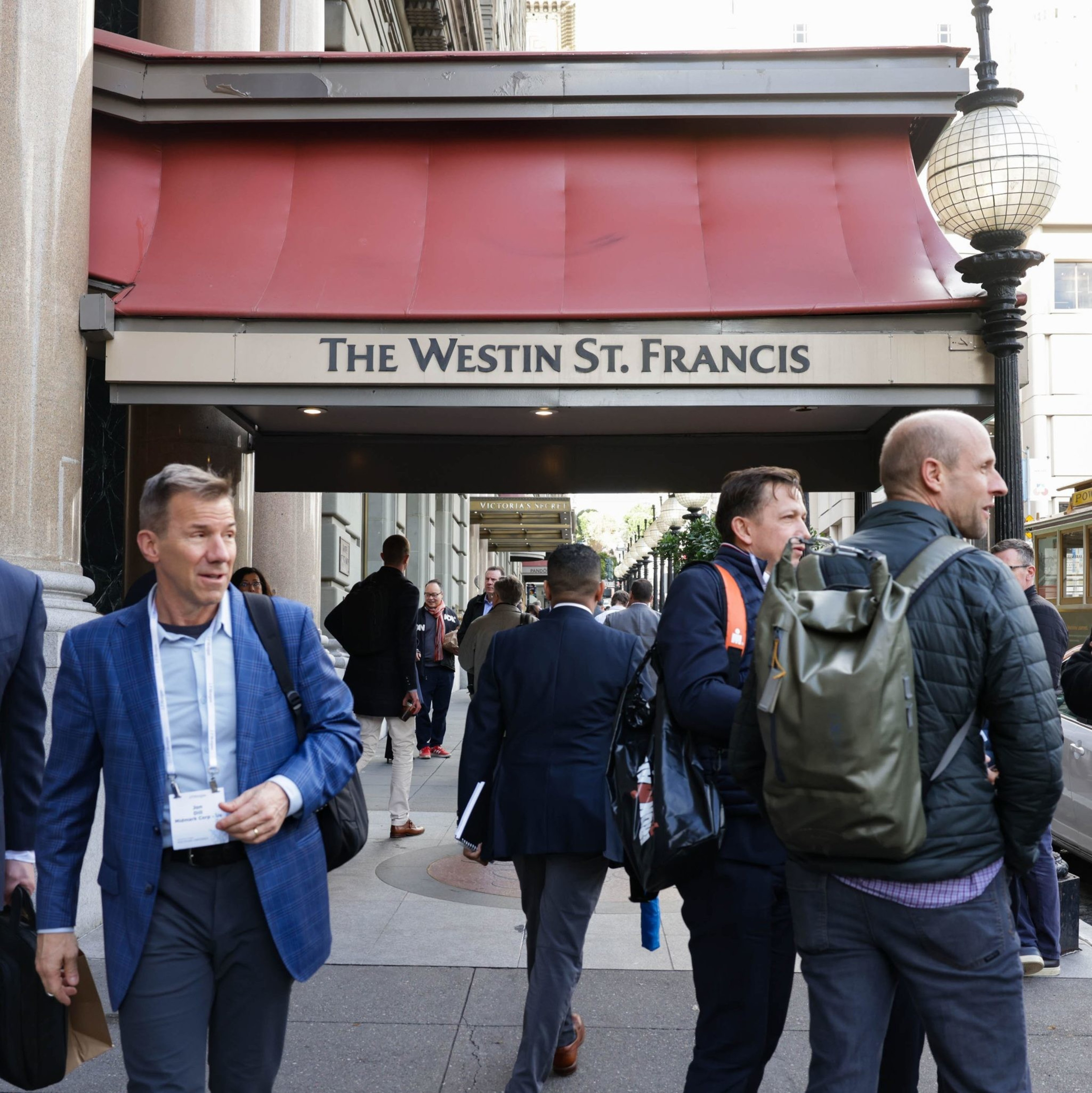 In front of The Westin St. Francis hotel, several professionally dressed men stand in conversation, while others walk by the red canopy entrance on a bustling sidewalk.