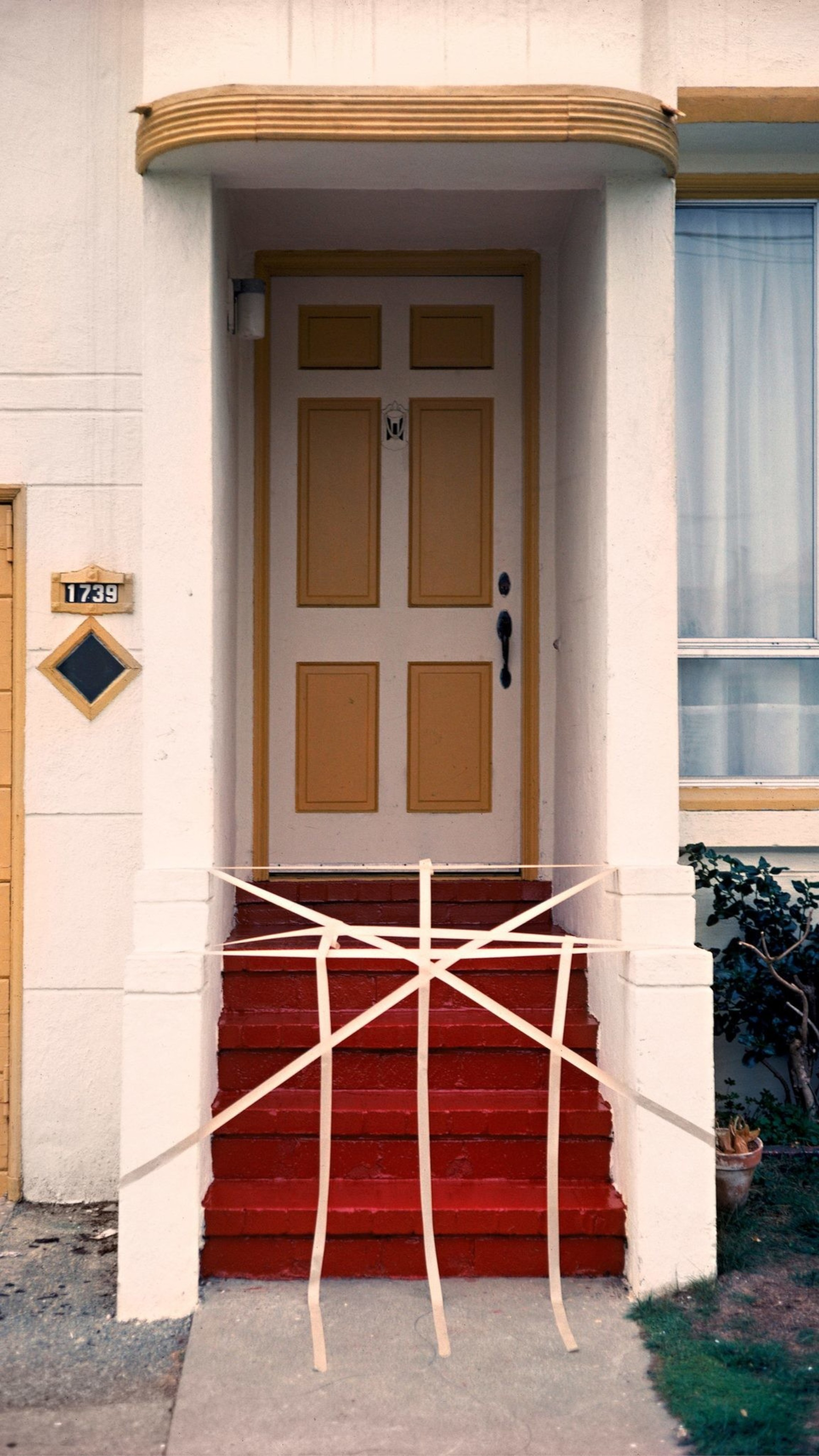 A red staircase leads to a tan, panelled door with white columns. The entrance is barricaded with white tape arranged in a web pattern.