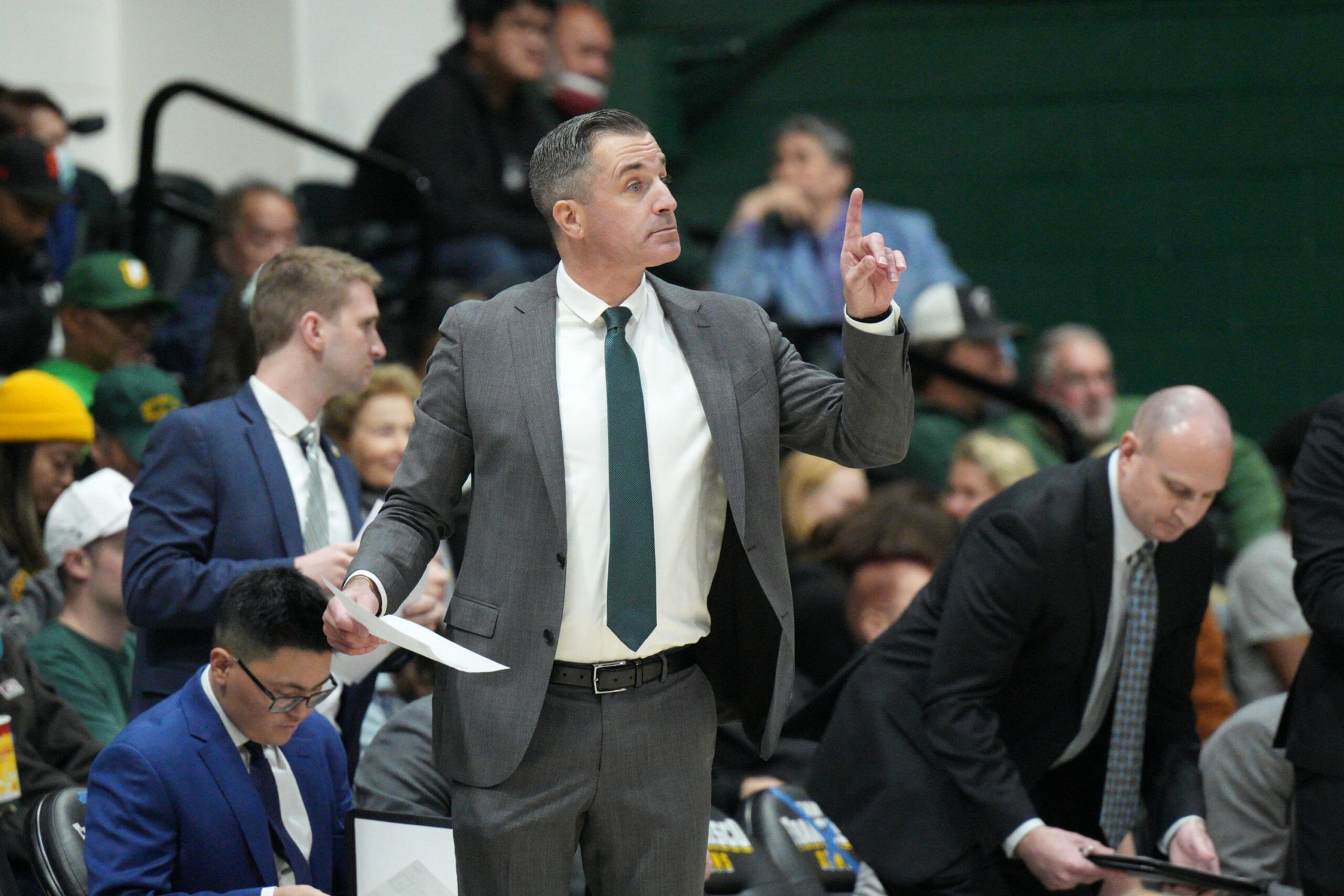 A man in a gray suit and green tie raises his index finger while holding papers, surrounded by other men in suits and spectators in a sports setting.