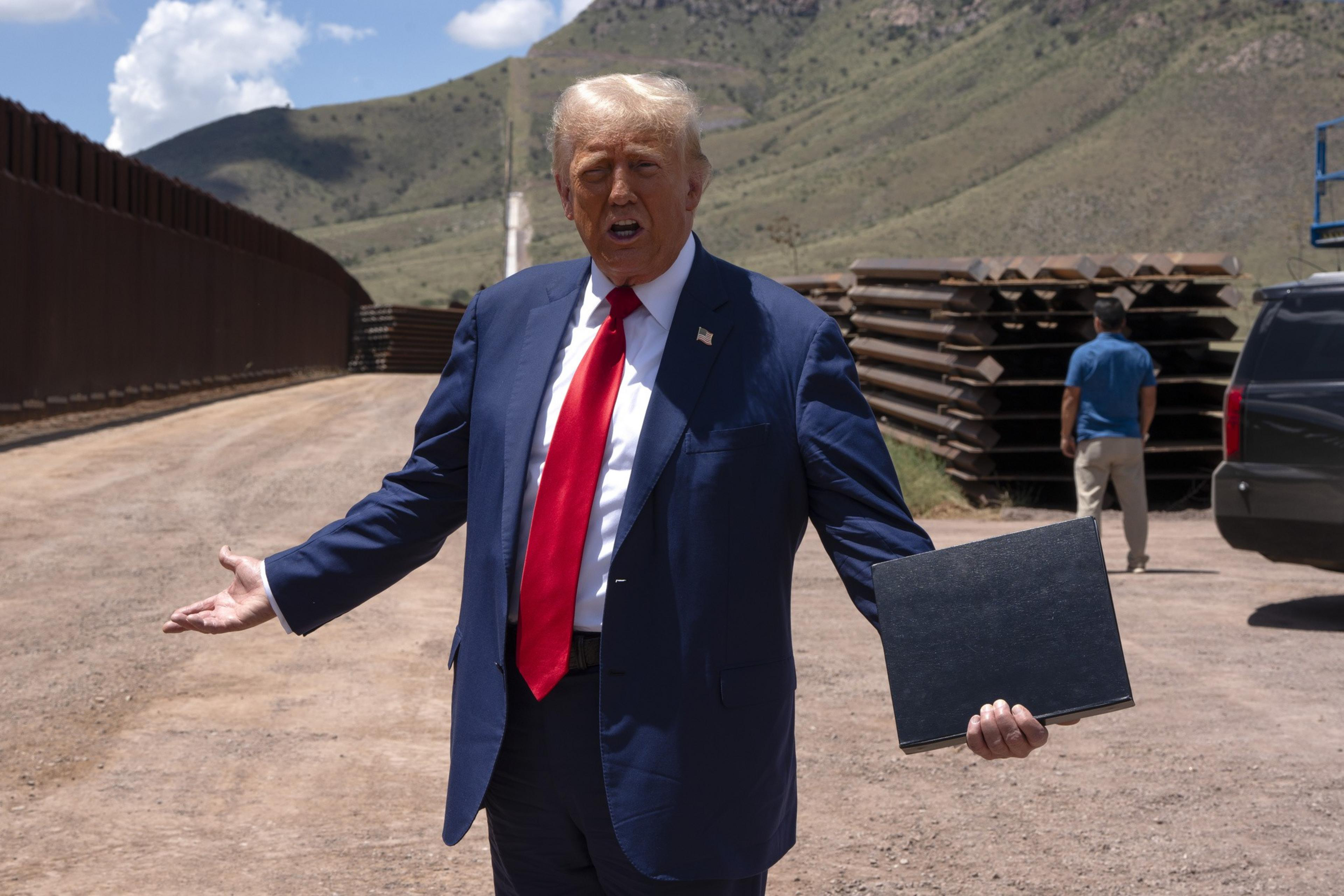 A man in a dark blue suit and red tie gestures with one hand while holding a folder in the other, standing near a border wall with mountains in the background.