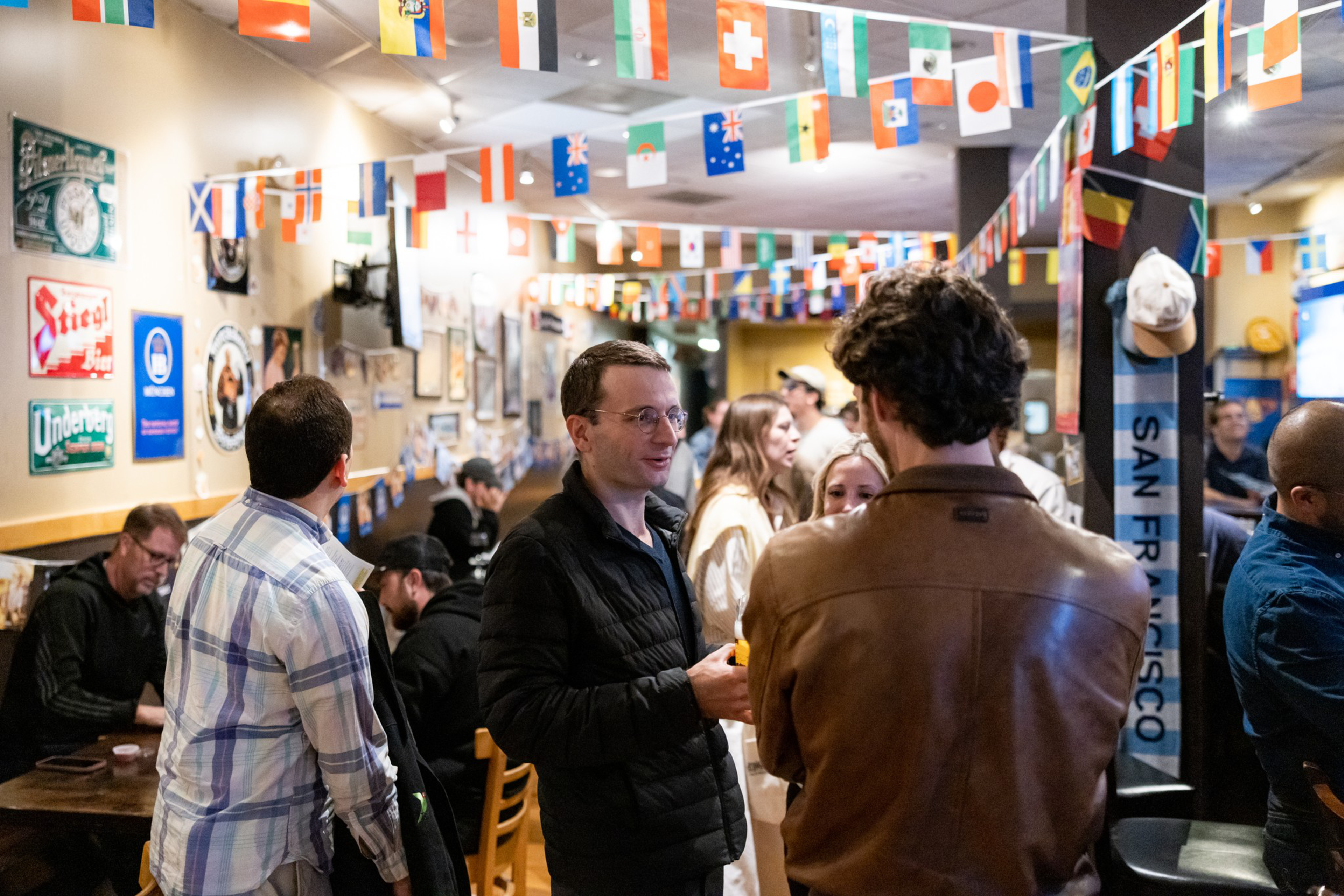 People are socializing in a lively bar decorated with international flags hanging from the ceiling and various signs on the walls.