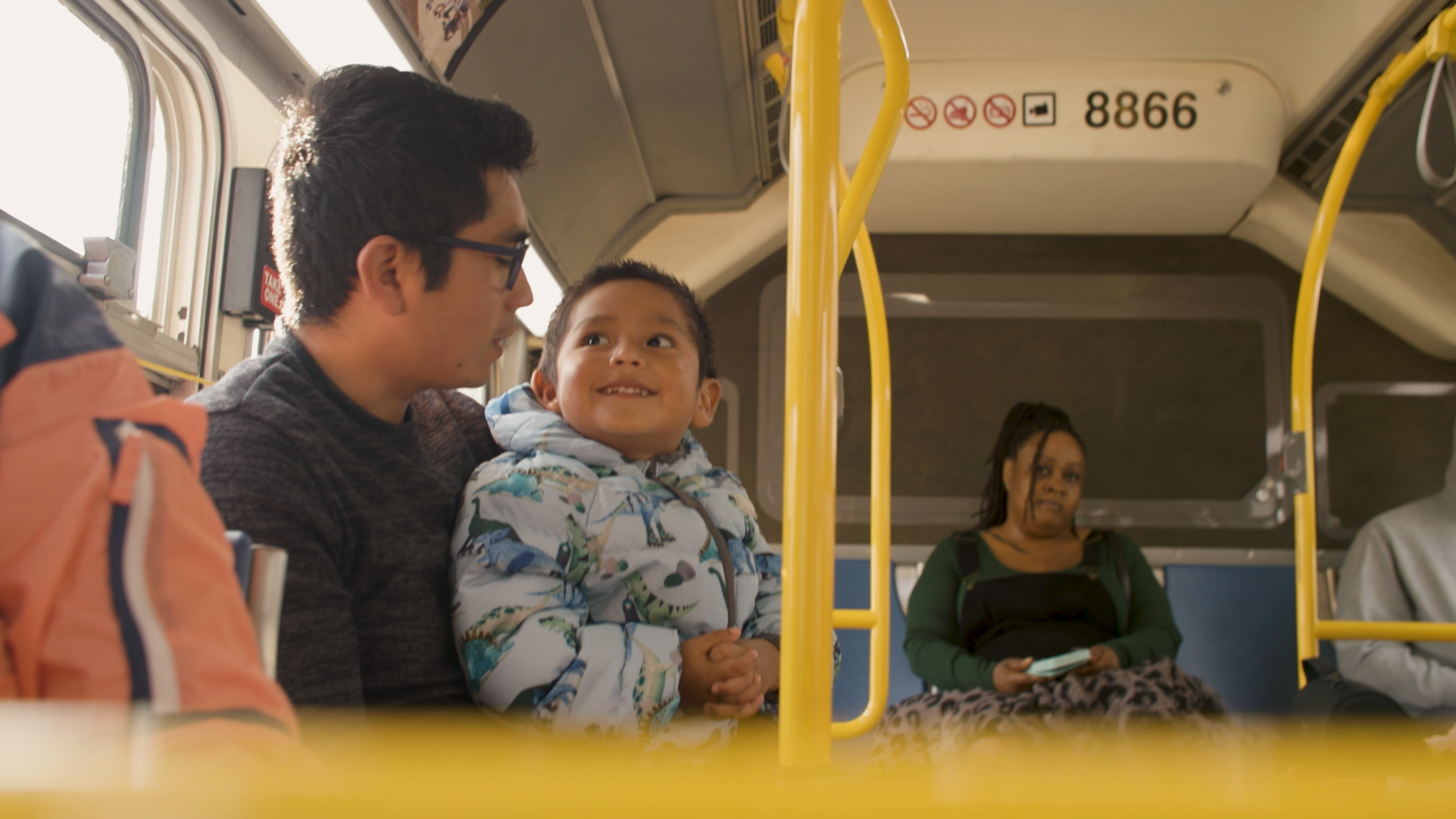 A man and child sit close together on a bus, the child smiling up at the man, while a woman and another person sit separately in the background.