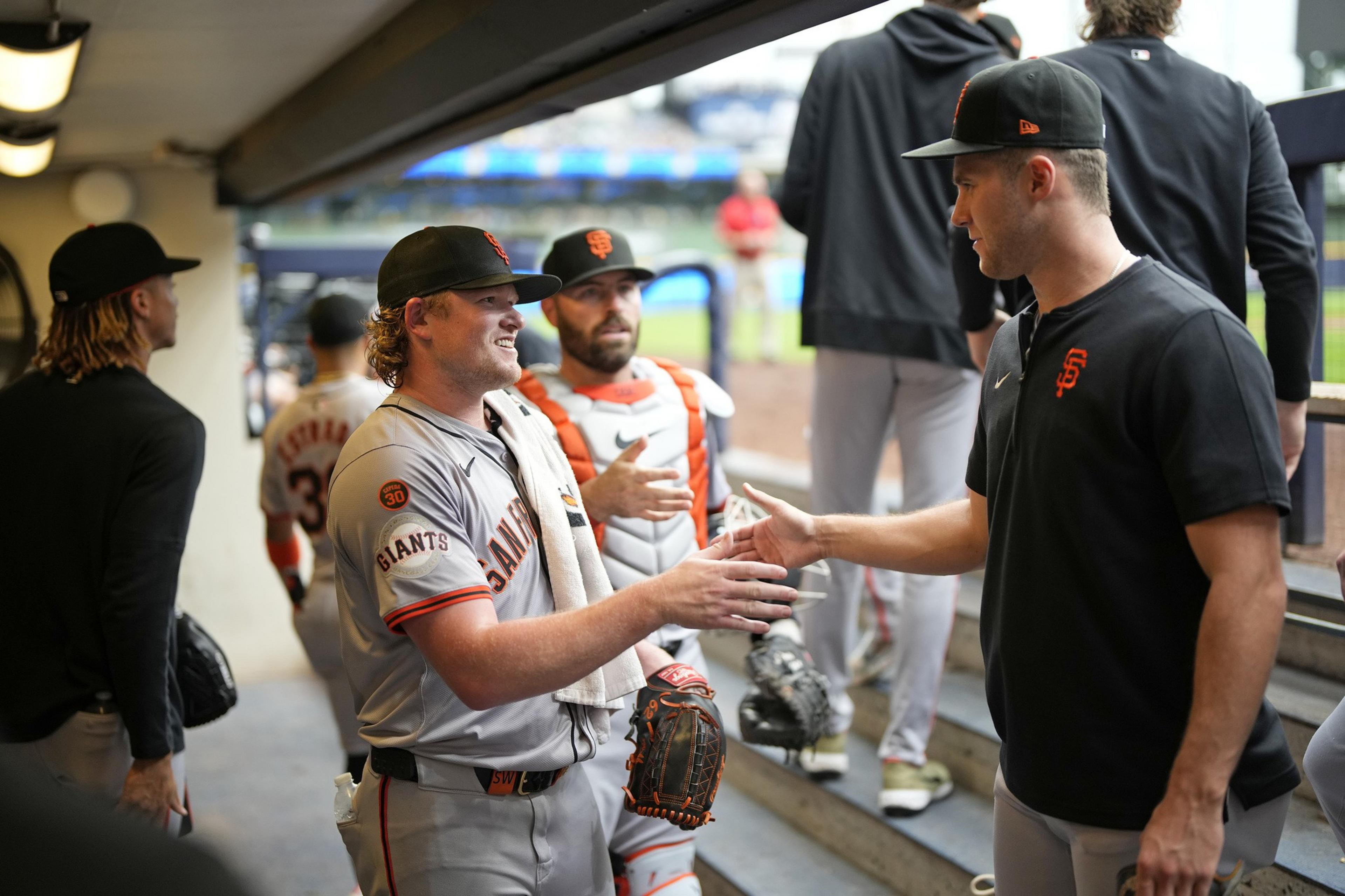 Two baseball players in Giants uniforms are shaking hands in a dugout, with more players in the background, one wearing catcher's gear.