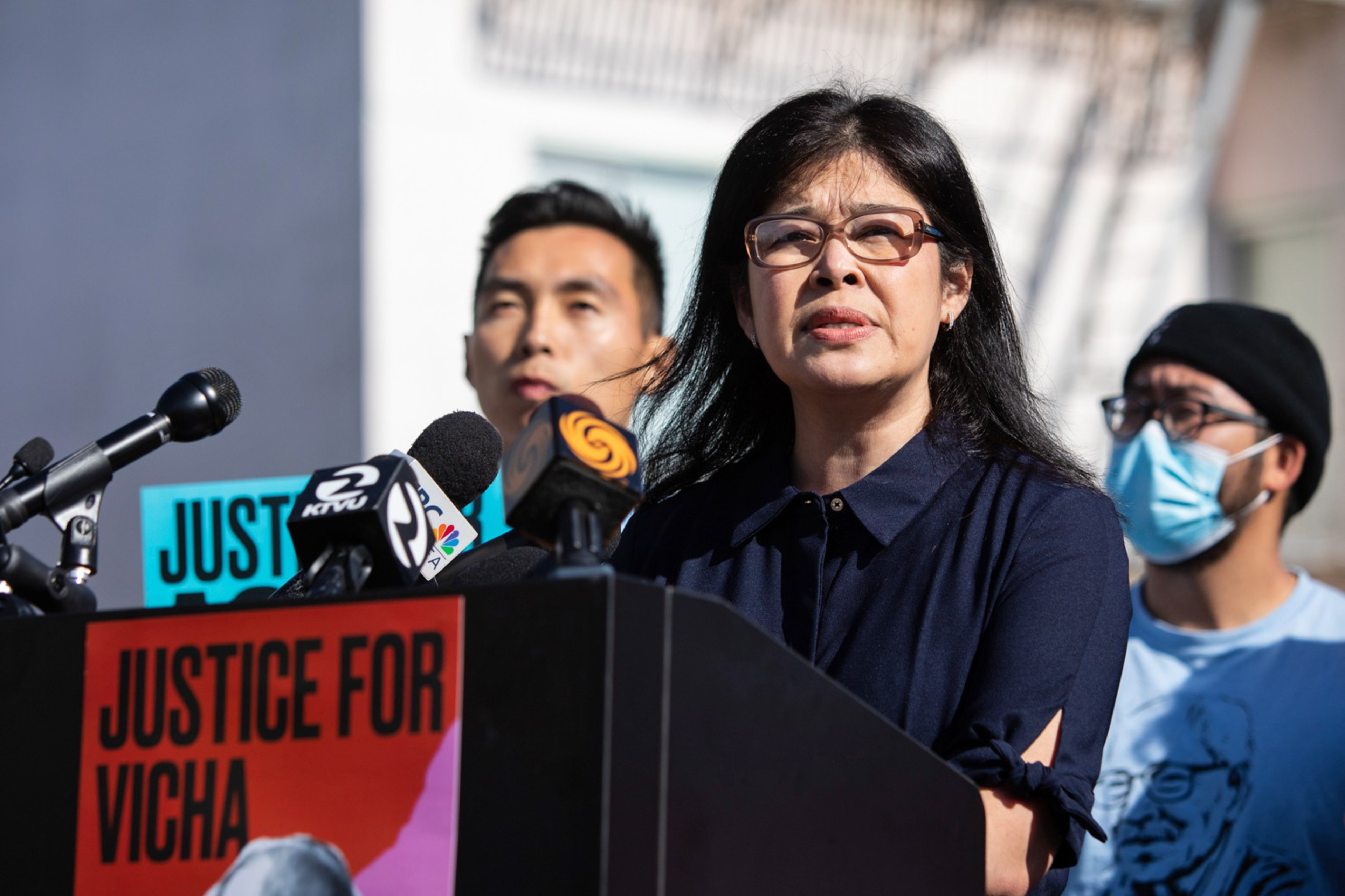 A woman speaks at a press conference with microphones, flanked by two men, one wearing a face mask, and a sign reading "Justice for Vicha."