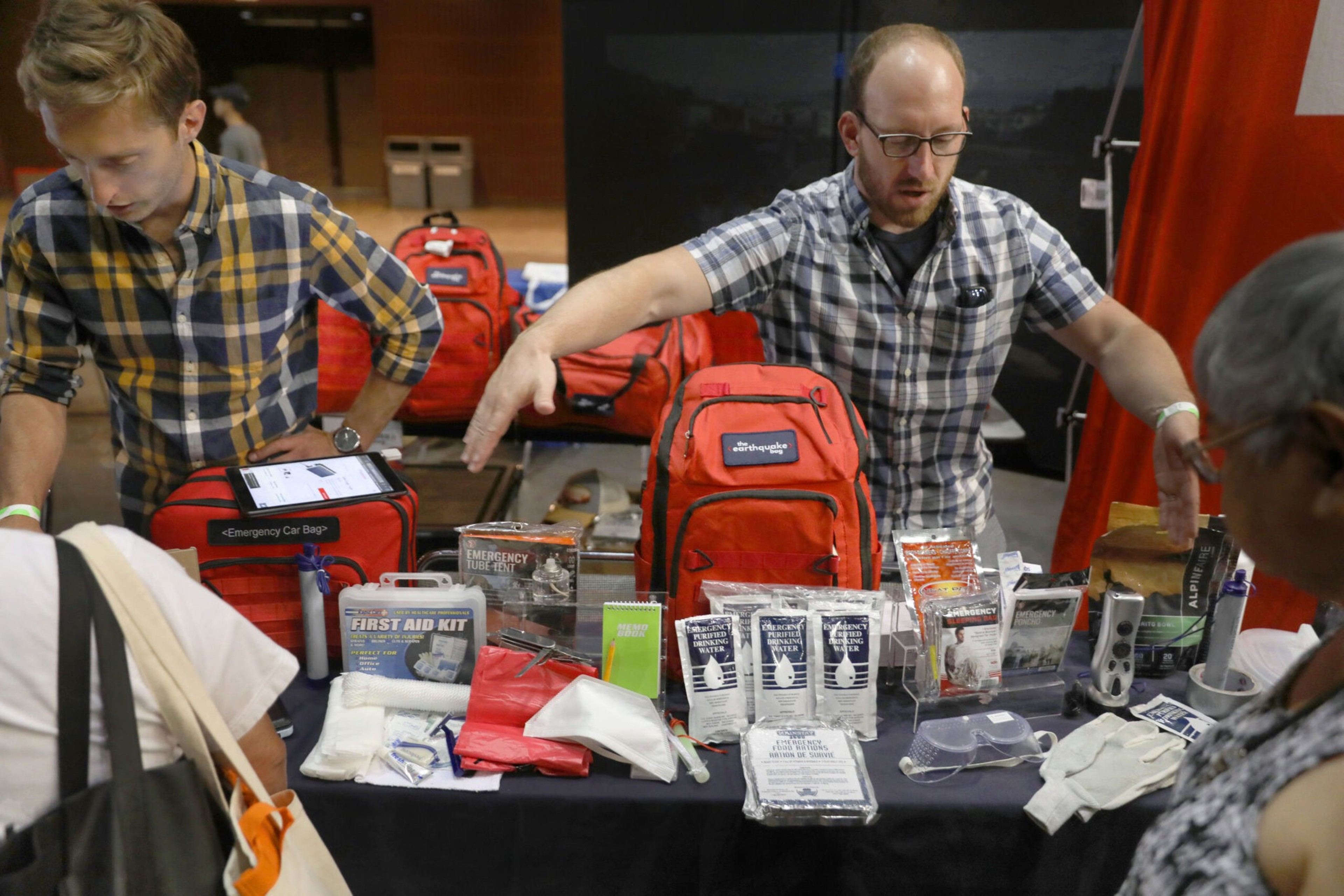 Two men in plaid shirts display a variety of emergency supplies, including first aid kits, gloves, water pouches, and red emergency bags on a table.