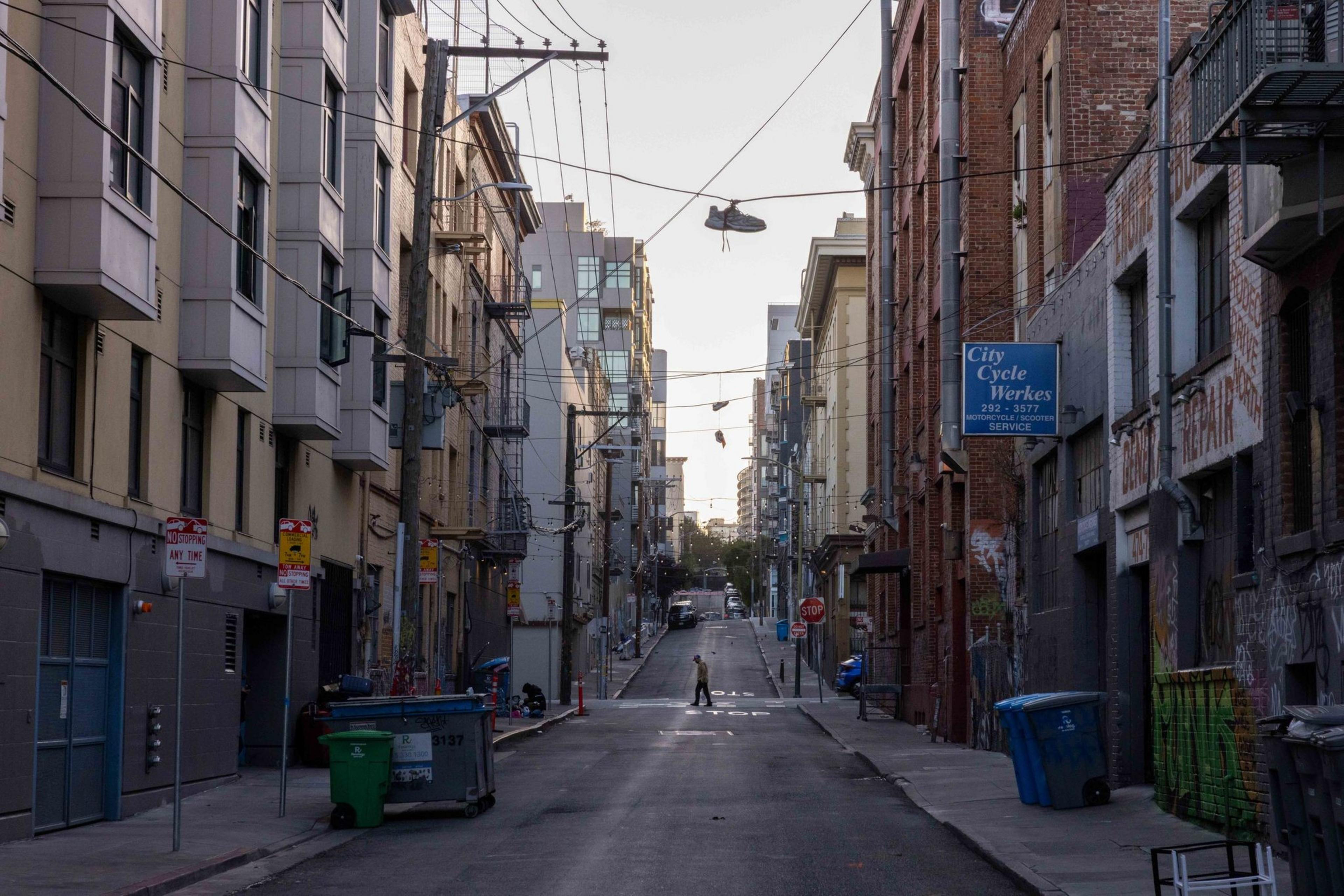 A narrow urban street lined with buildings has shoes hanging from overhead wires. A person stands in the distance, and various garbage bins are visible on the sidewalk.