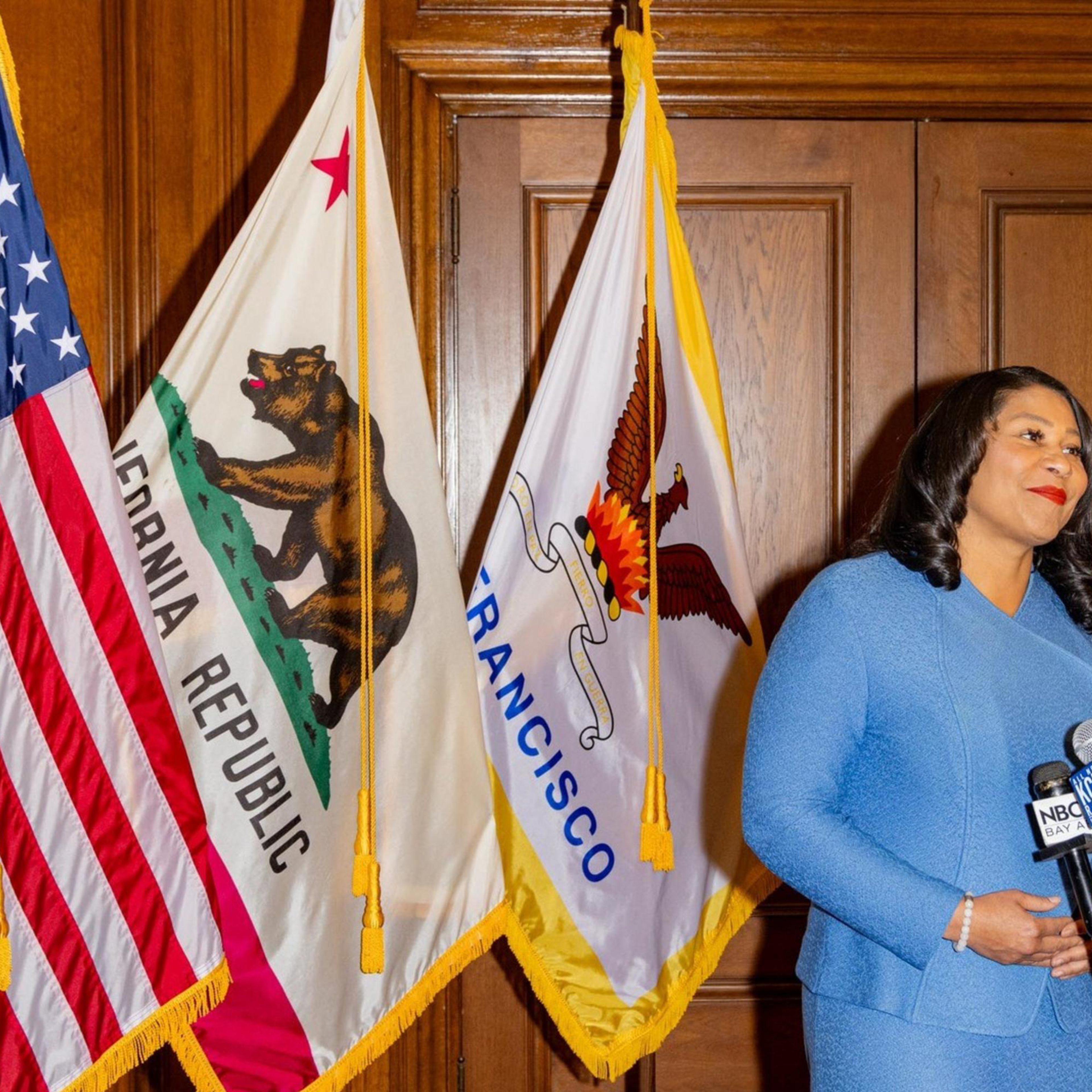 A person in a blue suit stands at a podium with multiple microphones, flanked by the U.S., California, and San Francisco flags inside a wood-paneled room.
