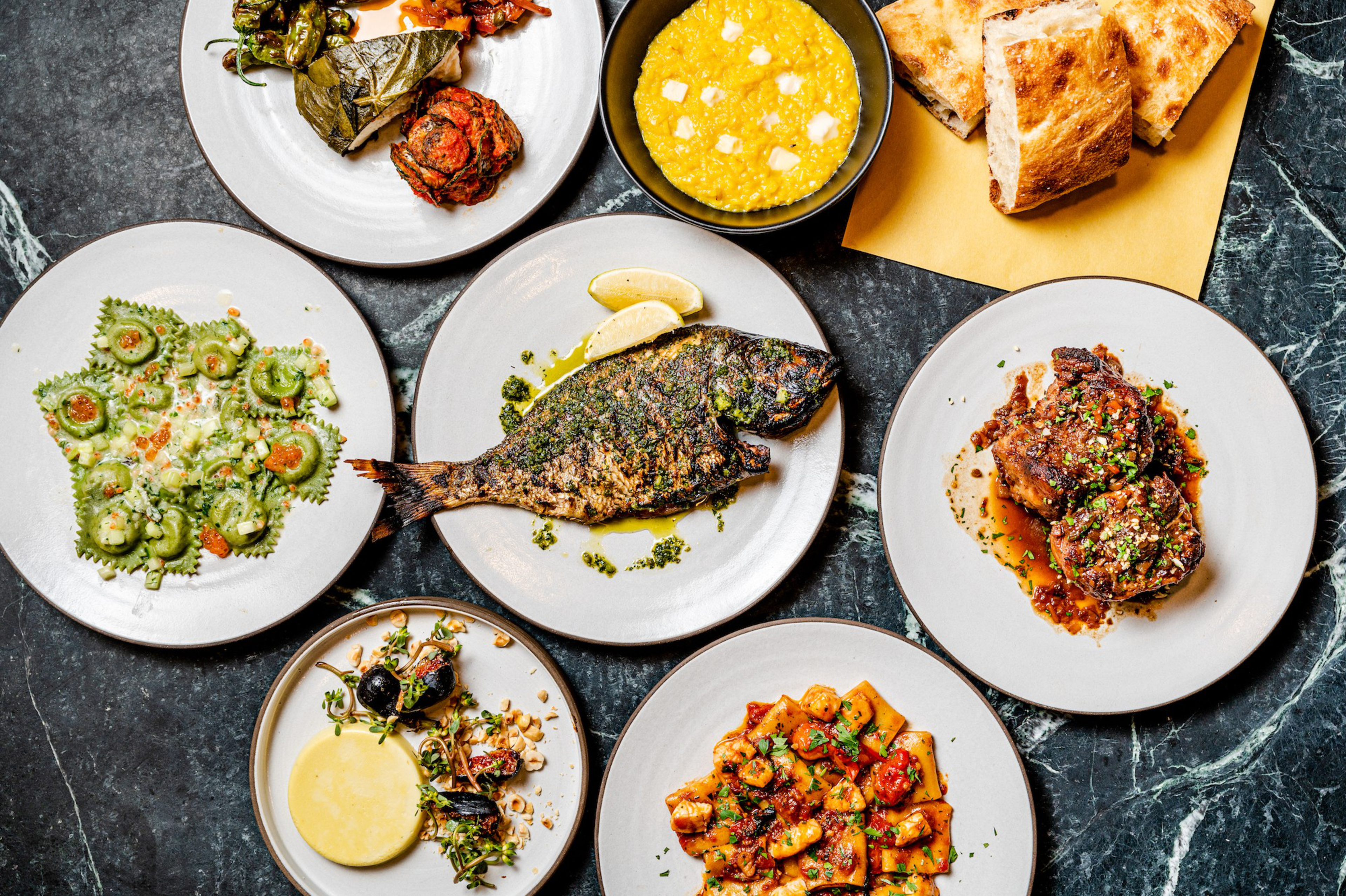 Grilled fish with herbs, polenta with cheese, flatbread, stuffed grape leaf, marinated meat, ravioli, cheese with olives, and a tomato-vegetable stew on plates.