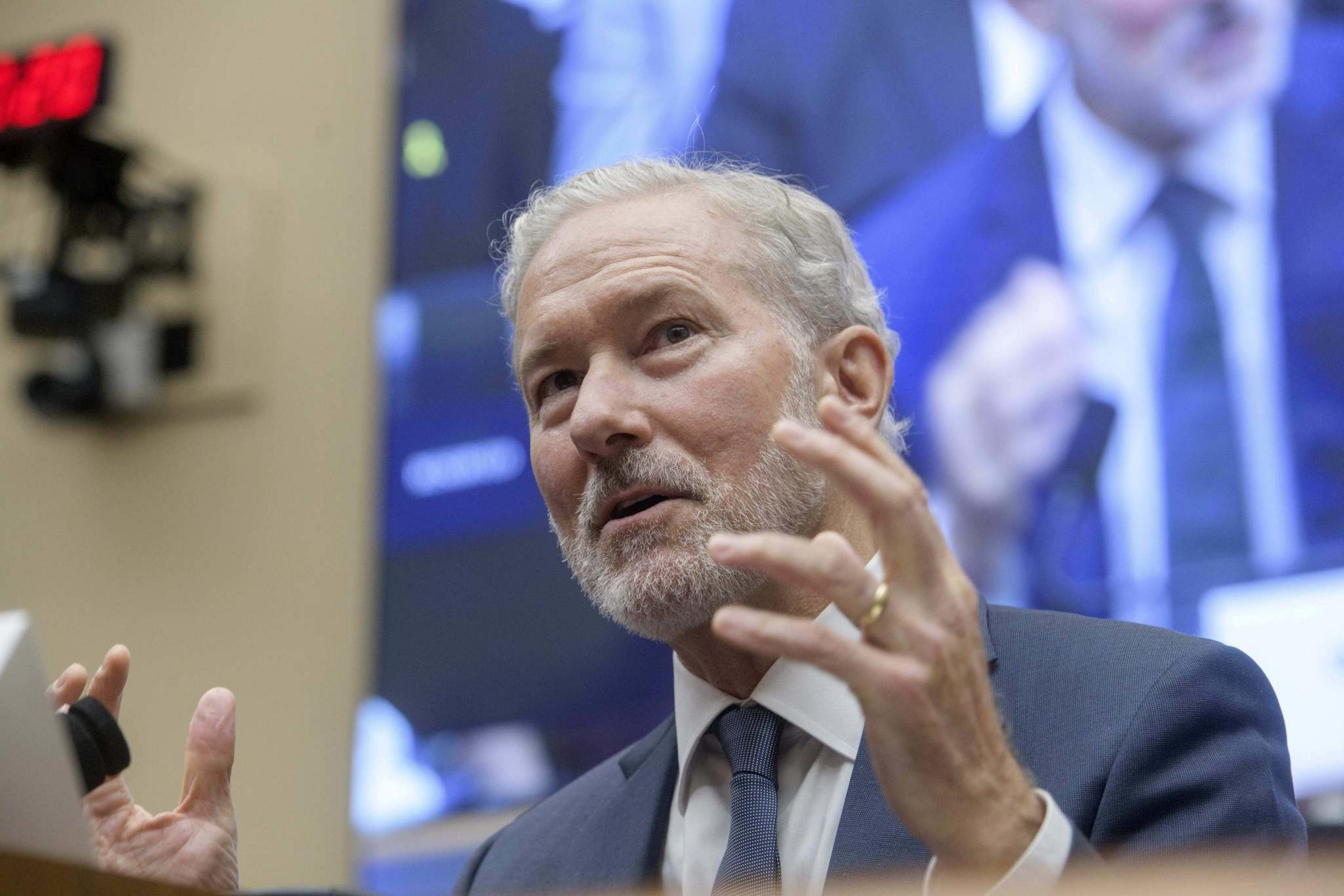 A man with light-colored hair and beard, wearing a suit and tie, gestures with his hands while speaking in front of a blurred background screen.