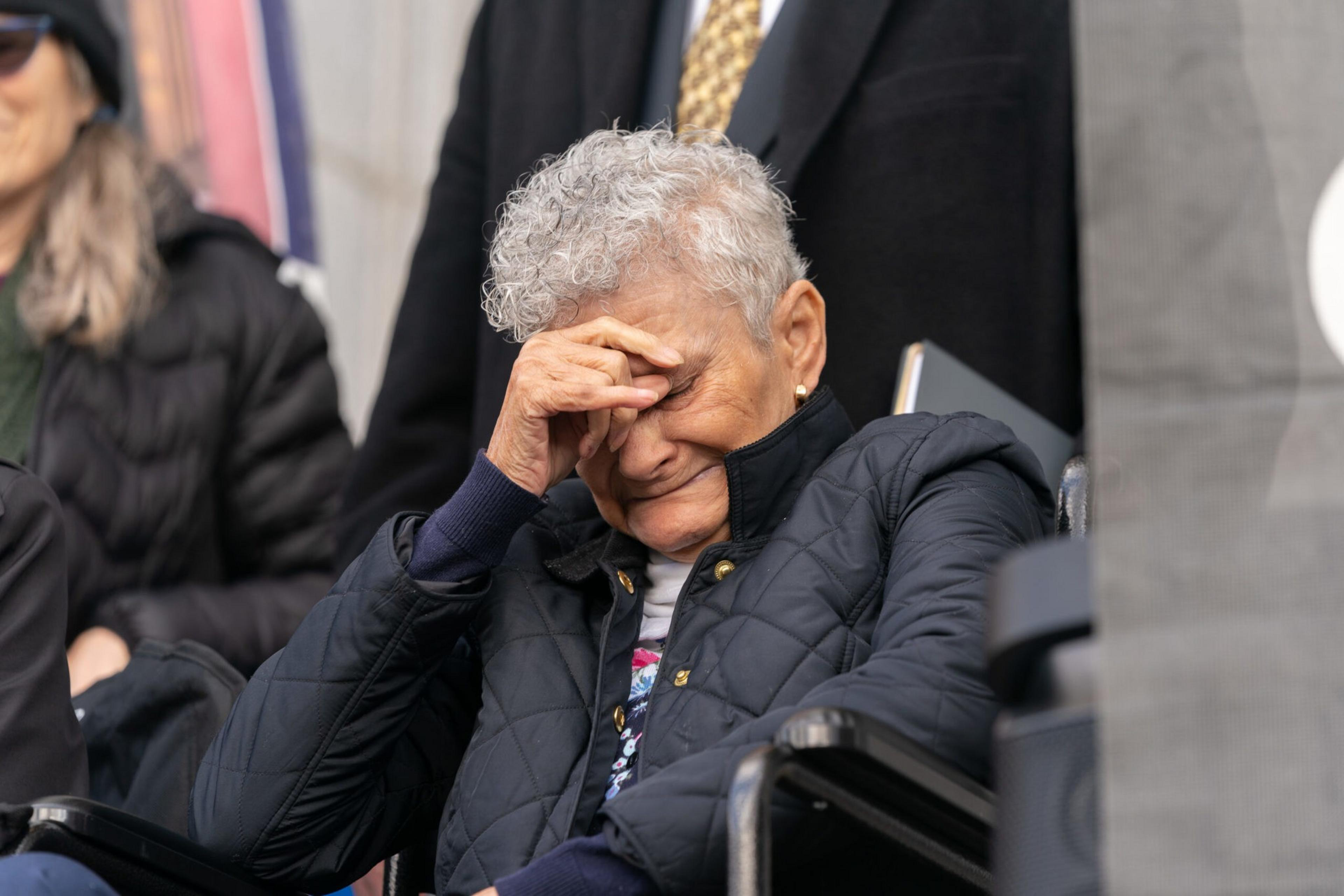 An elderly person with gray curly hair sits in a wheelchair, wearing a dark jacket, eyes closed, and hand covering their forehead in apparent distress.