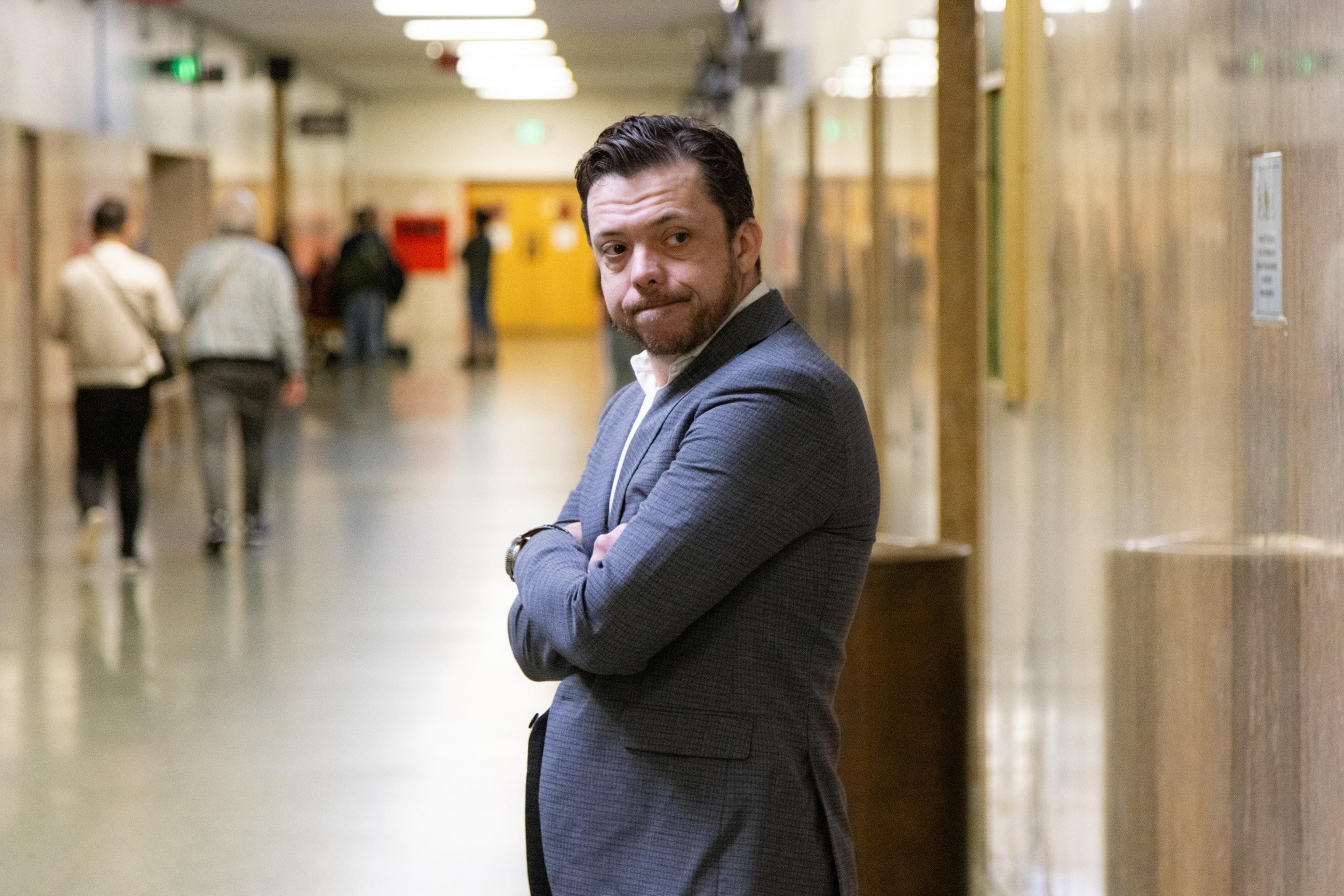 A man in a gray suit stands in a hallway with crossed arms, looking to the side. The hallway is busy with people walking in the background.