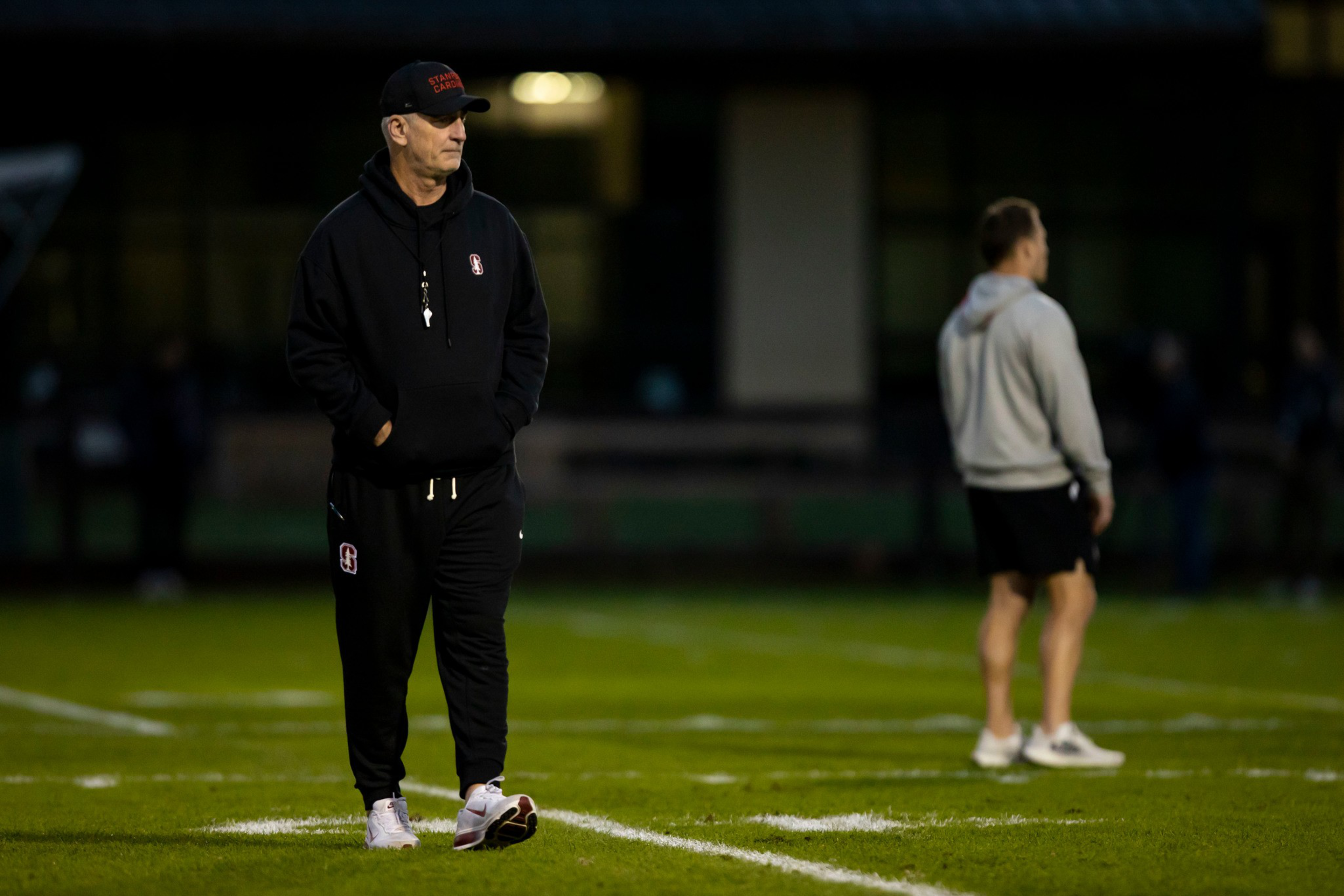 A man in black Stanford sportswear and a cap stands on a football field with hands in pockets, while another person in gray and black stands blurred in the background.