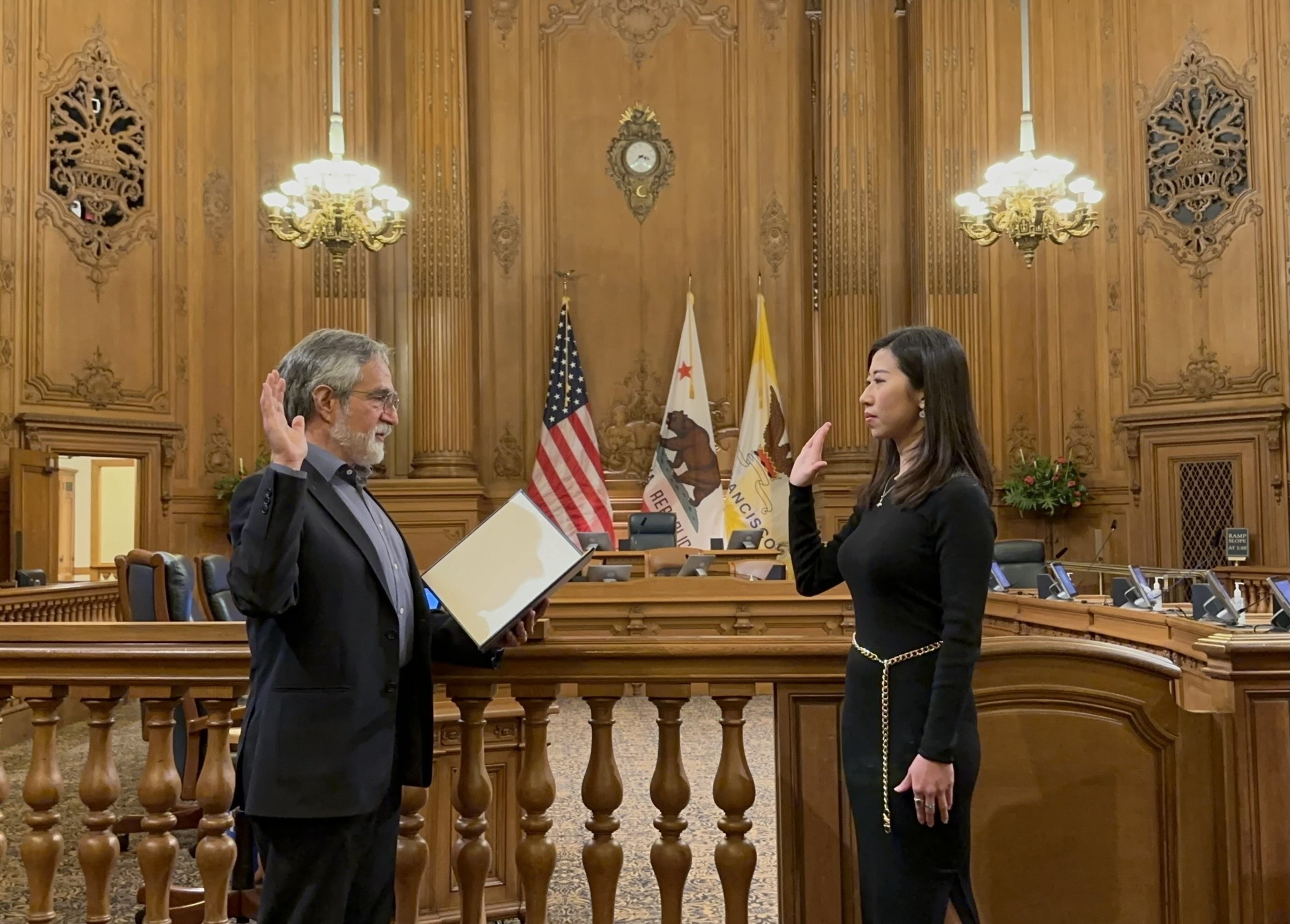 An oath-taking ceremony in a wood-paneled room with two people raising their right hands, an American flag, and ornate dÃ©cor.