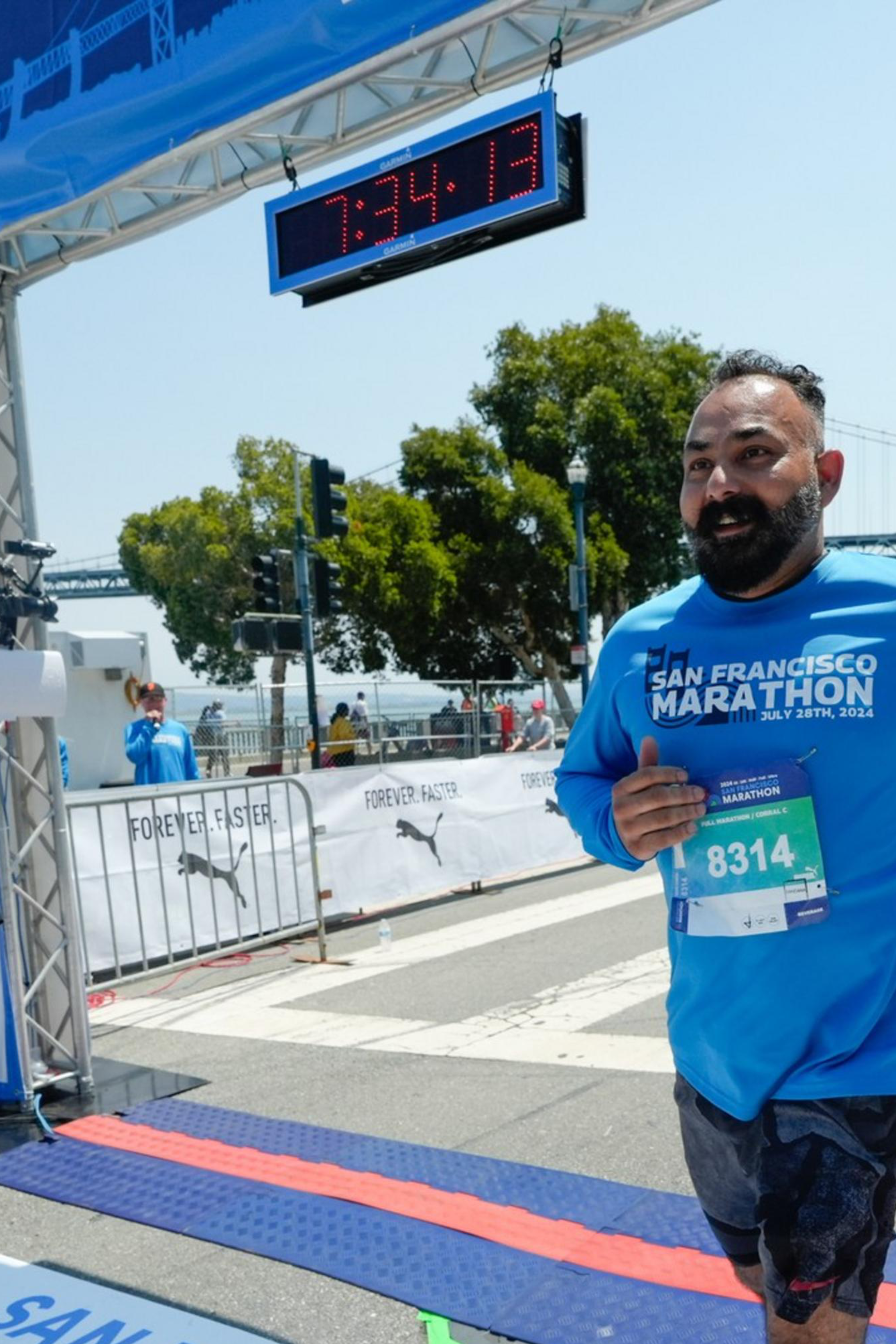 A man in a blue &quot;San Francisco Marathon&quot; shirt crosses the marathon finish line, with a digital clock above and a bridge in the background.