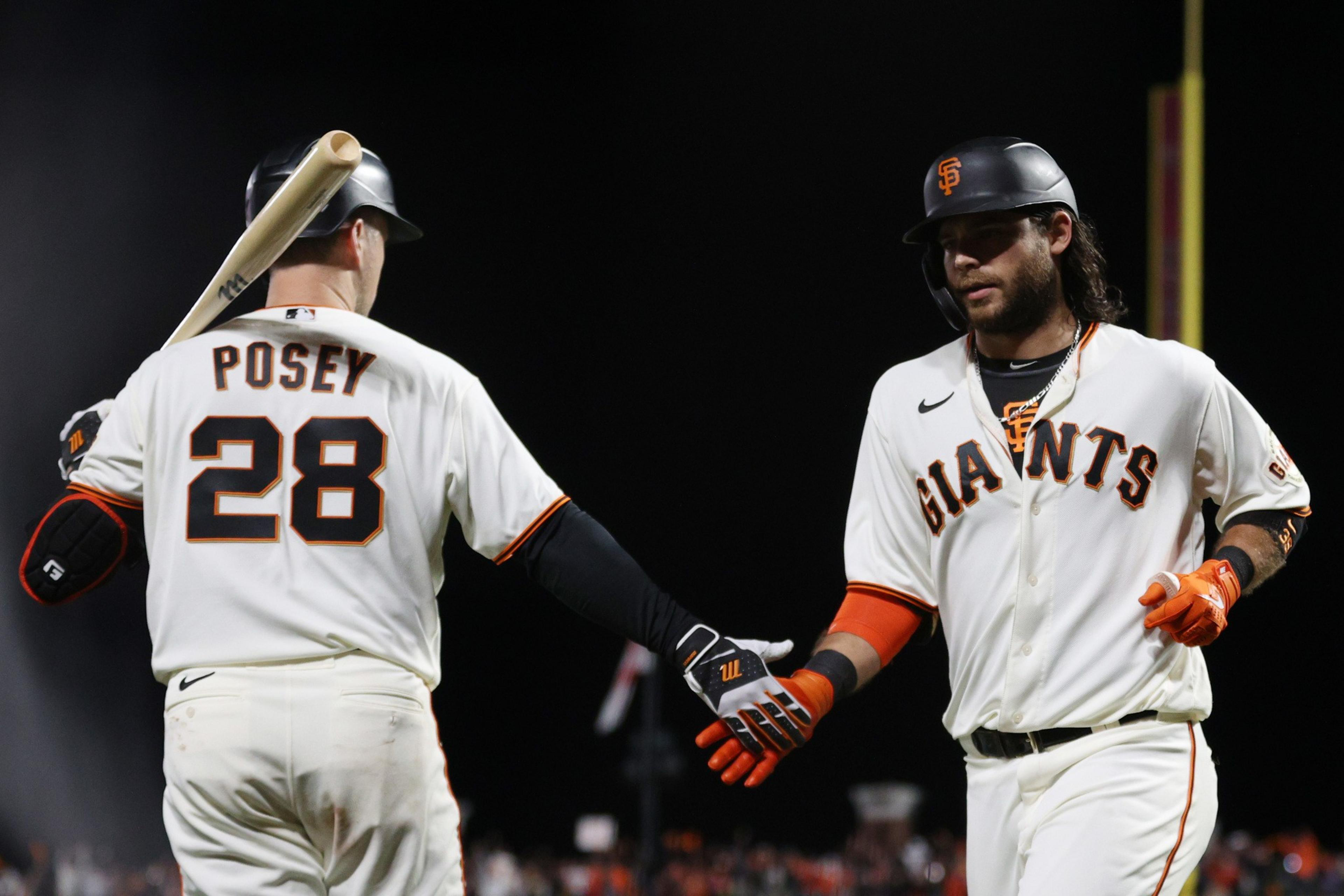 Two baseball players in white jerseys high-five on the field at night. One is holding a bat, and their uniforms say &quot;Giants.&quot; The scene feels celebratory.
