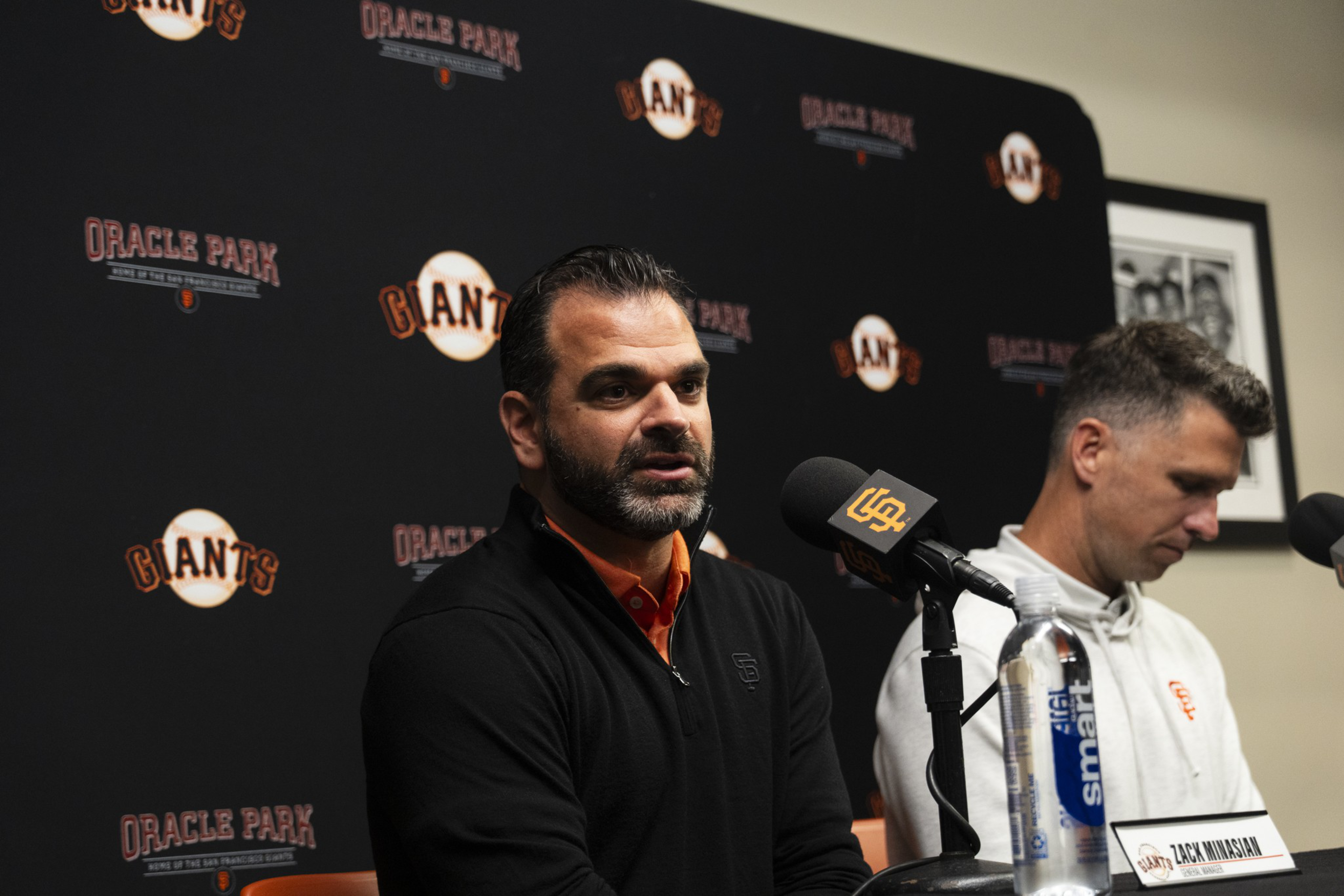 A man in a black jacket speaks into a microphone with a Giants logo backdrop, while another man in a white jacket sits beside him looking down.