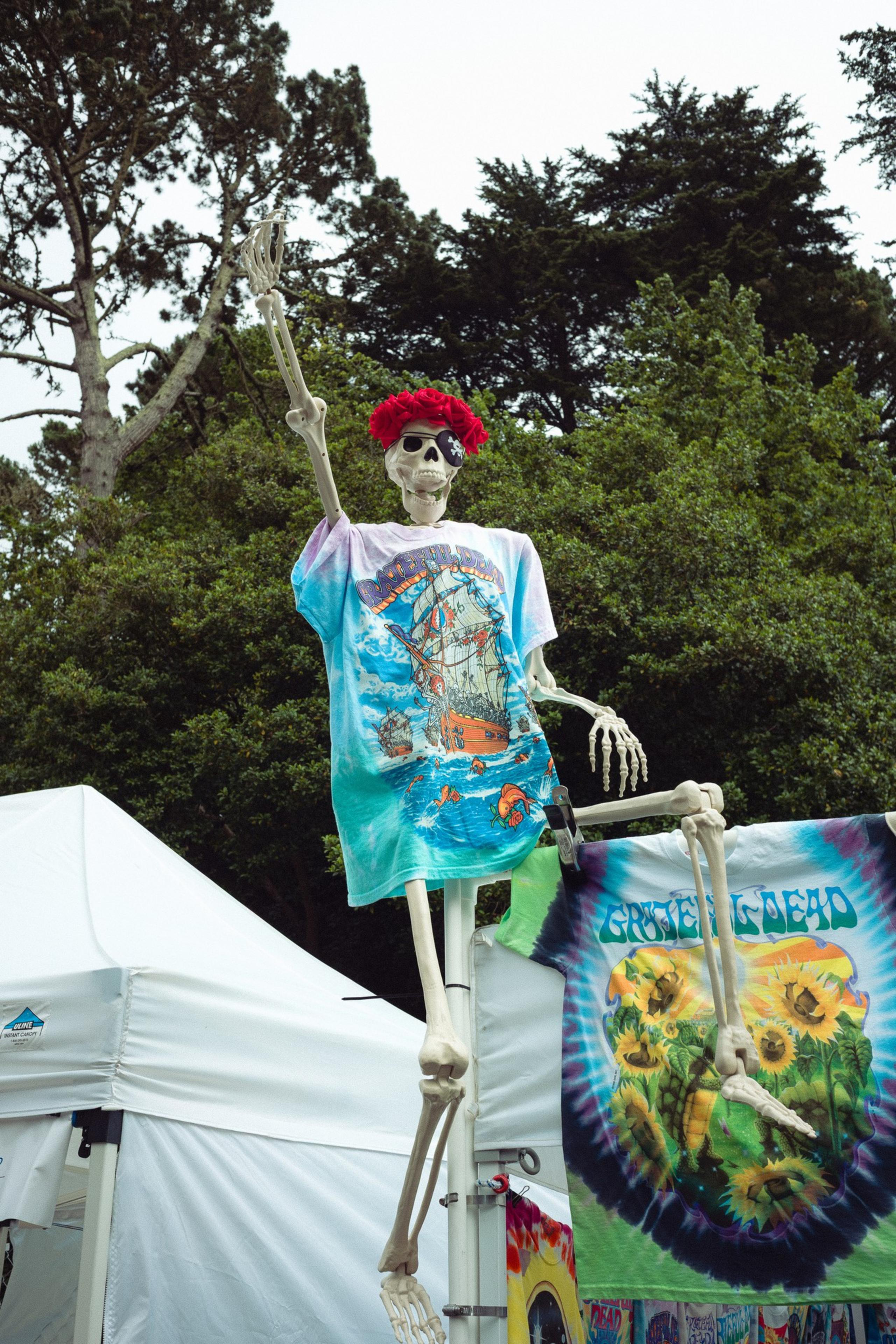 A skeleton wearing a red flower crown and a colorful ship-themed shirt sits on a pole, waving its hand, with tie-dye shirts hanging nearby.