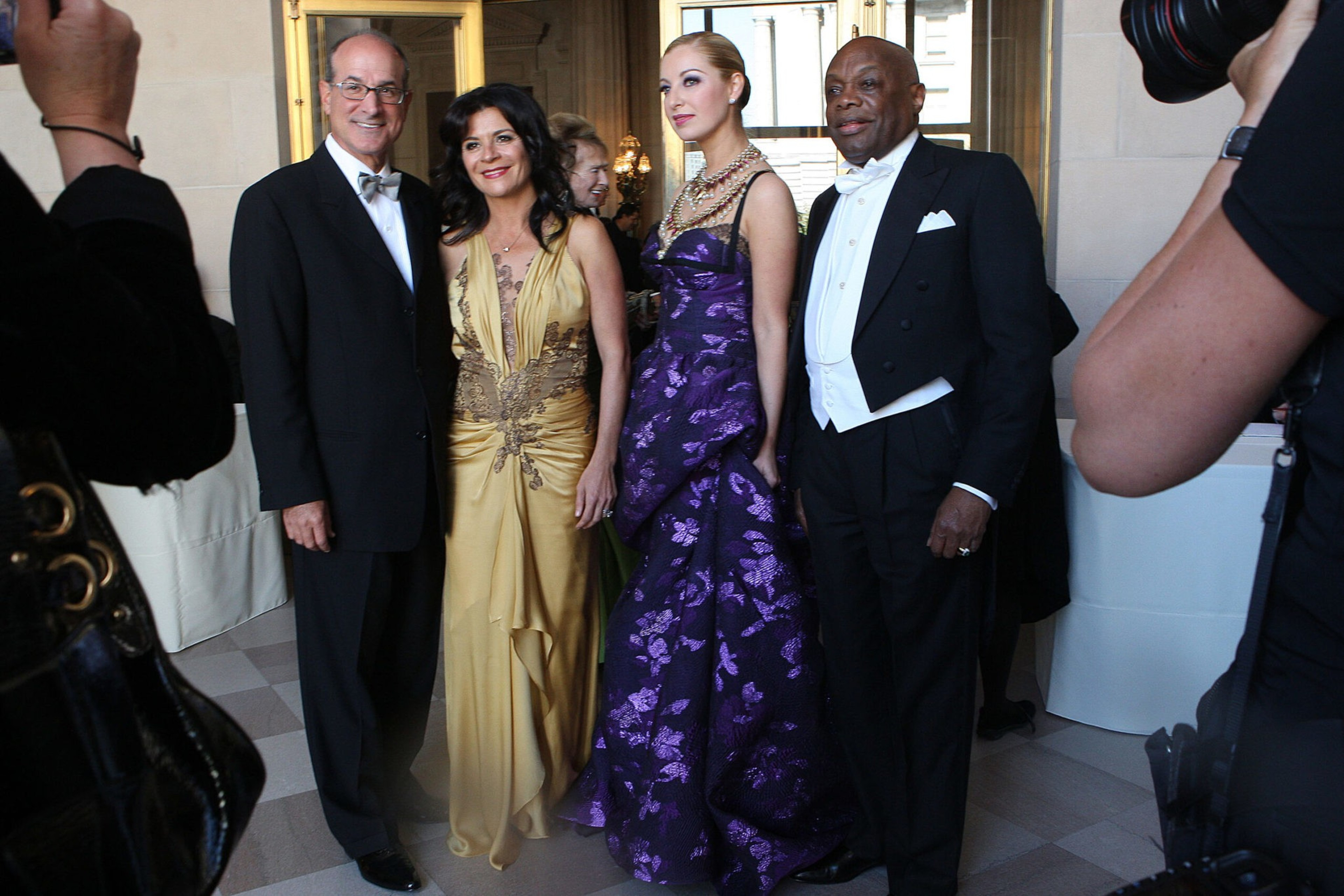 Two men in tuxedos and two women in elegant gowns—one gold, one purple—pose together indoors while photographers capture the moment.
