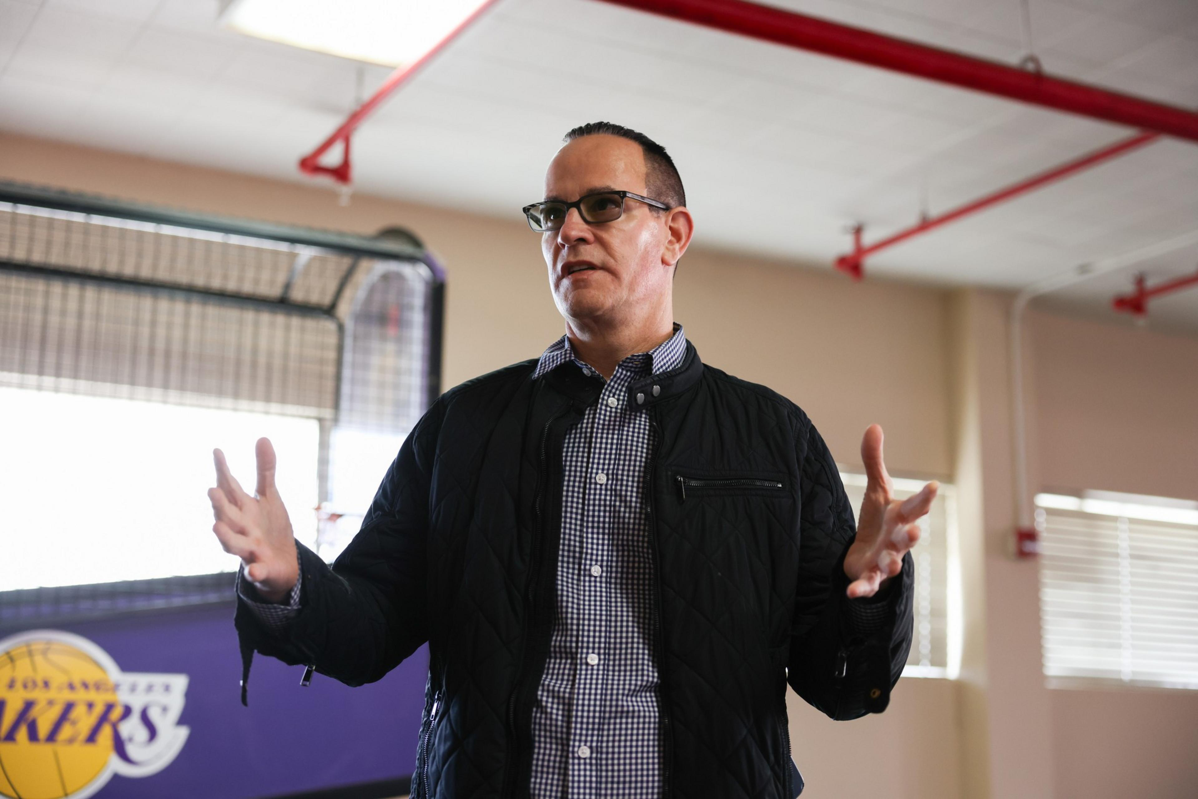 A man wearing glasses, a checkered shirt, and a dark jacket gestures with his hands while standing indoors near a basketball-themed backdrop.